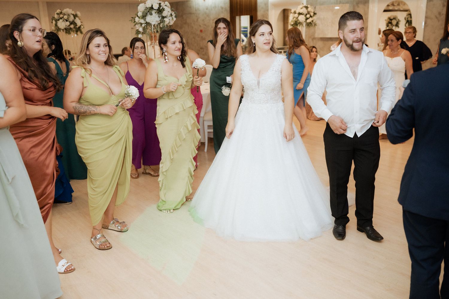 Wedding party and guests stand together in an elegant ballroom with polished wood flooring for a group photo.