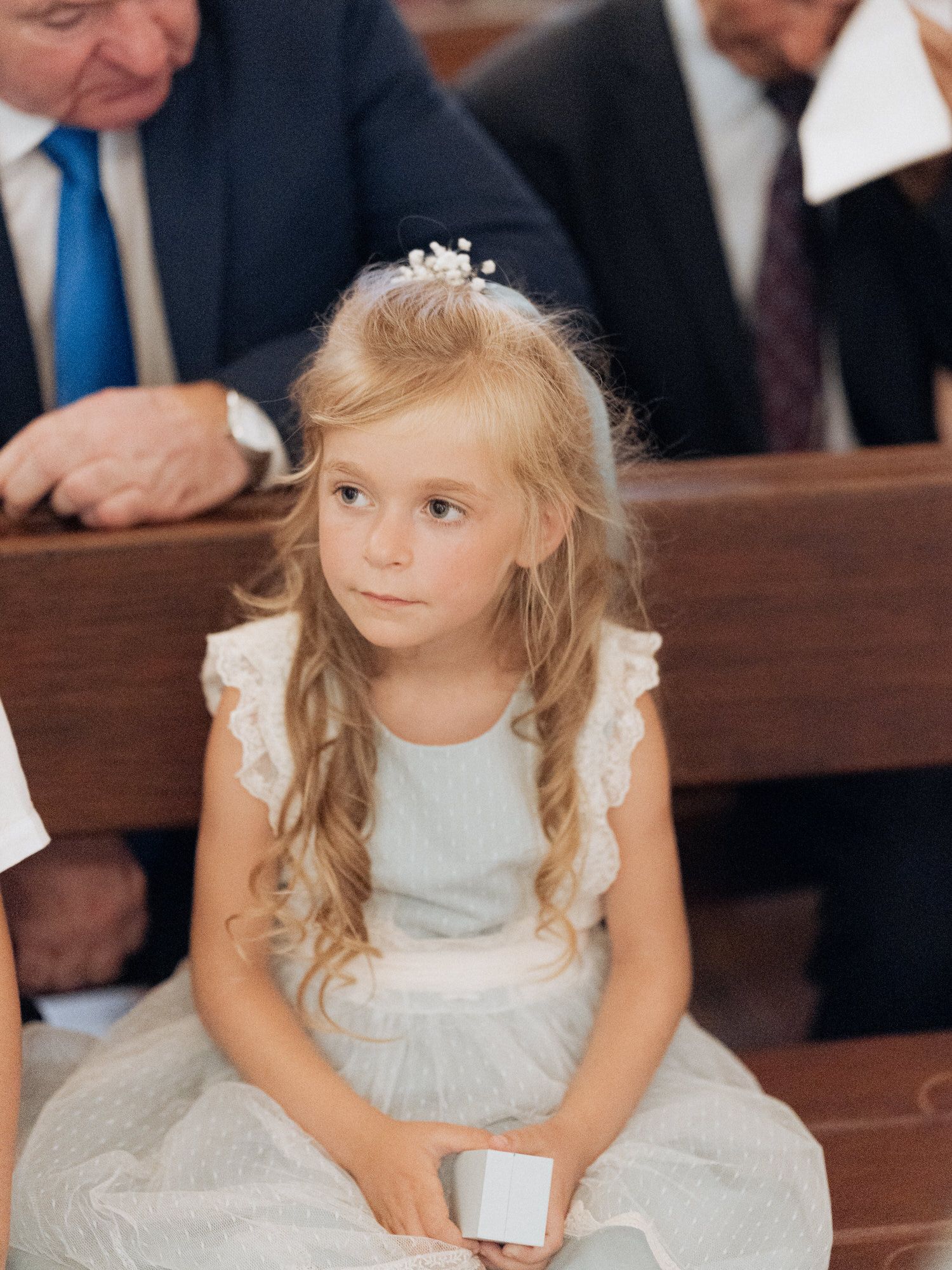 A young flower girl in a white lace dress sits on a wooden church pew during a wedding ceremony.