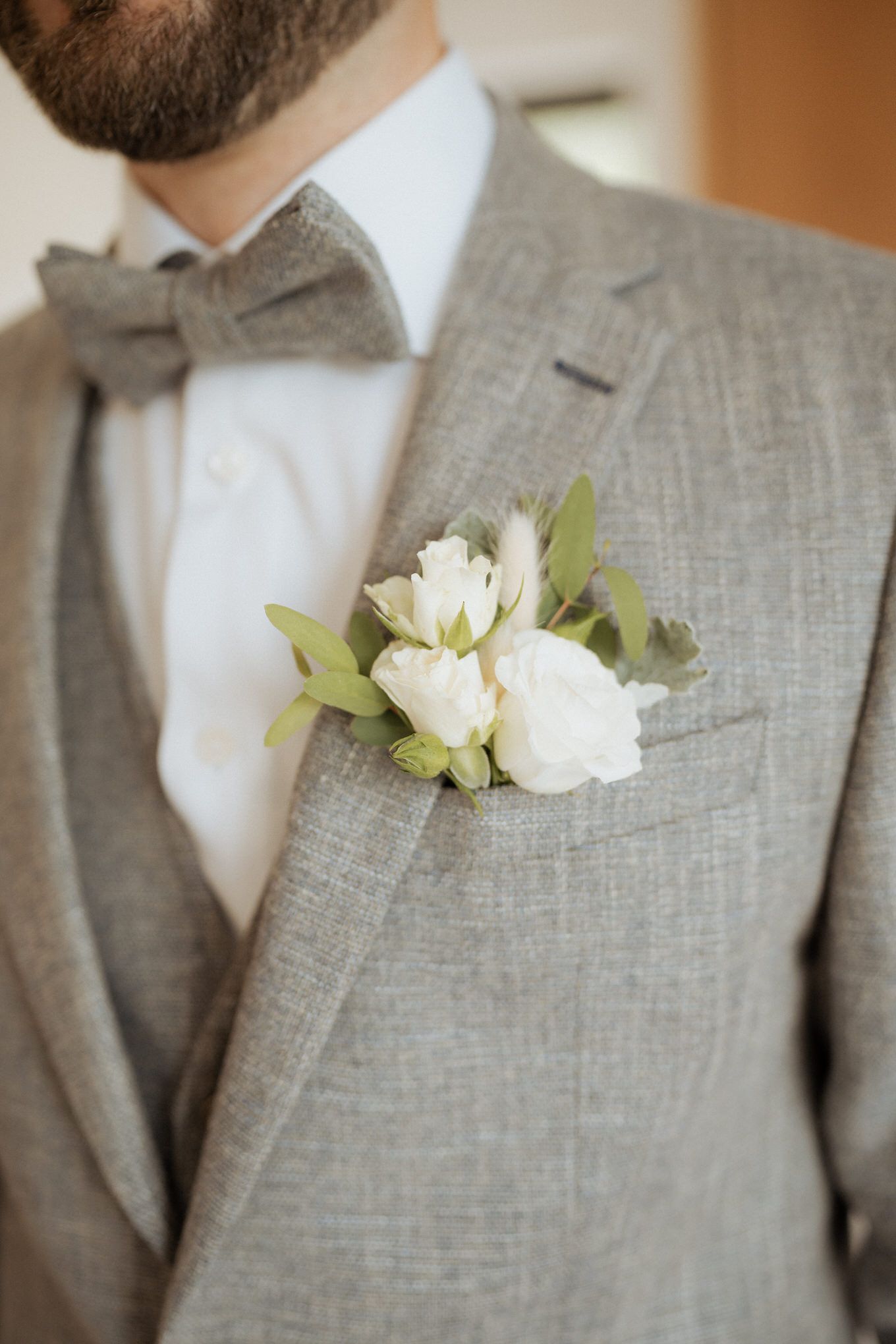 Close-up of a white floral boutonniere with green leaves pinned to a light gray linen suit lapel.