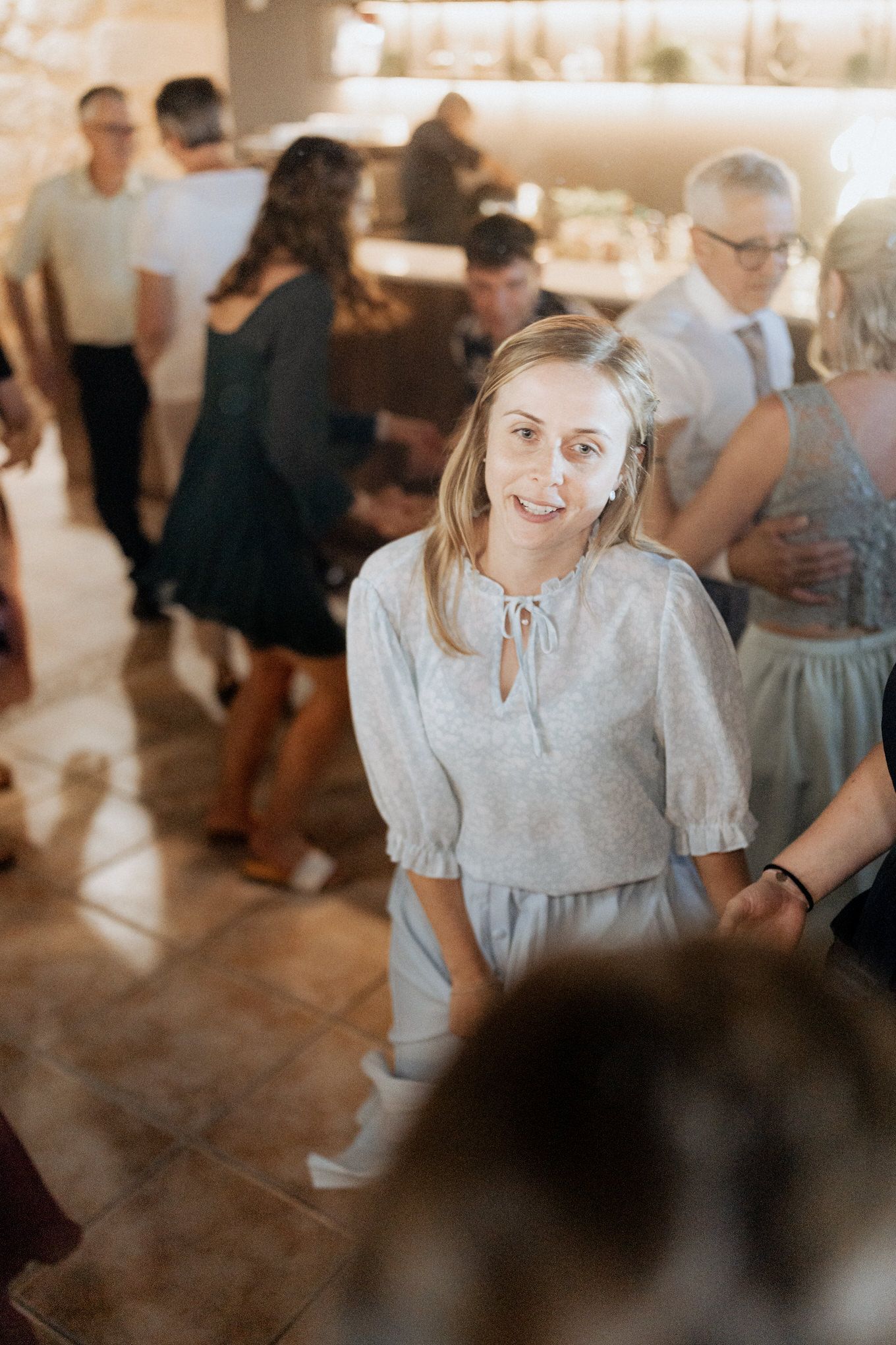 A person wearing a light gray puffed sleeve dress stands smiling at a social gathering with others mingling in the background.