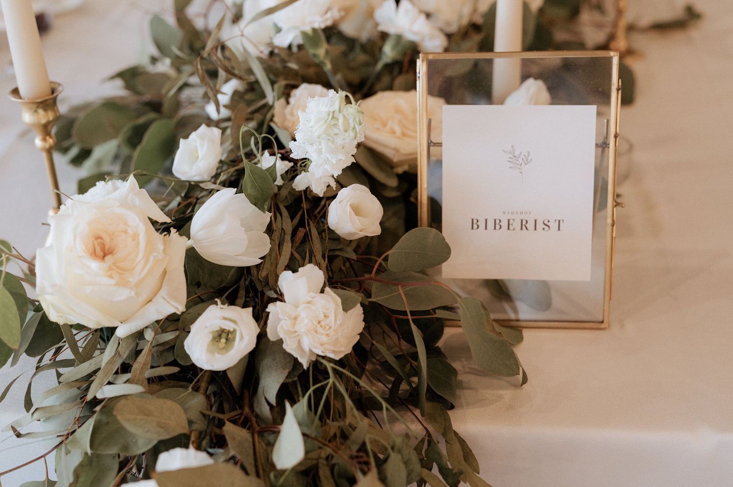Close-up of white rose and eucalyptus floral arrangement with directional signage in a gold frame.