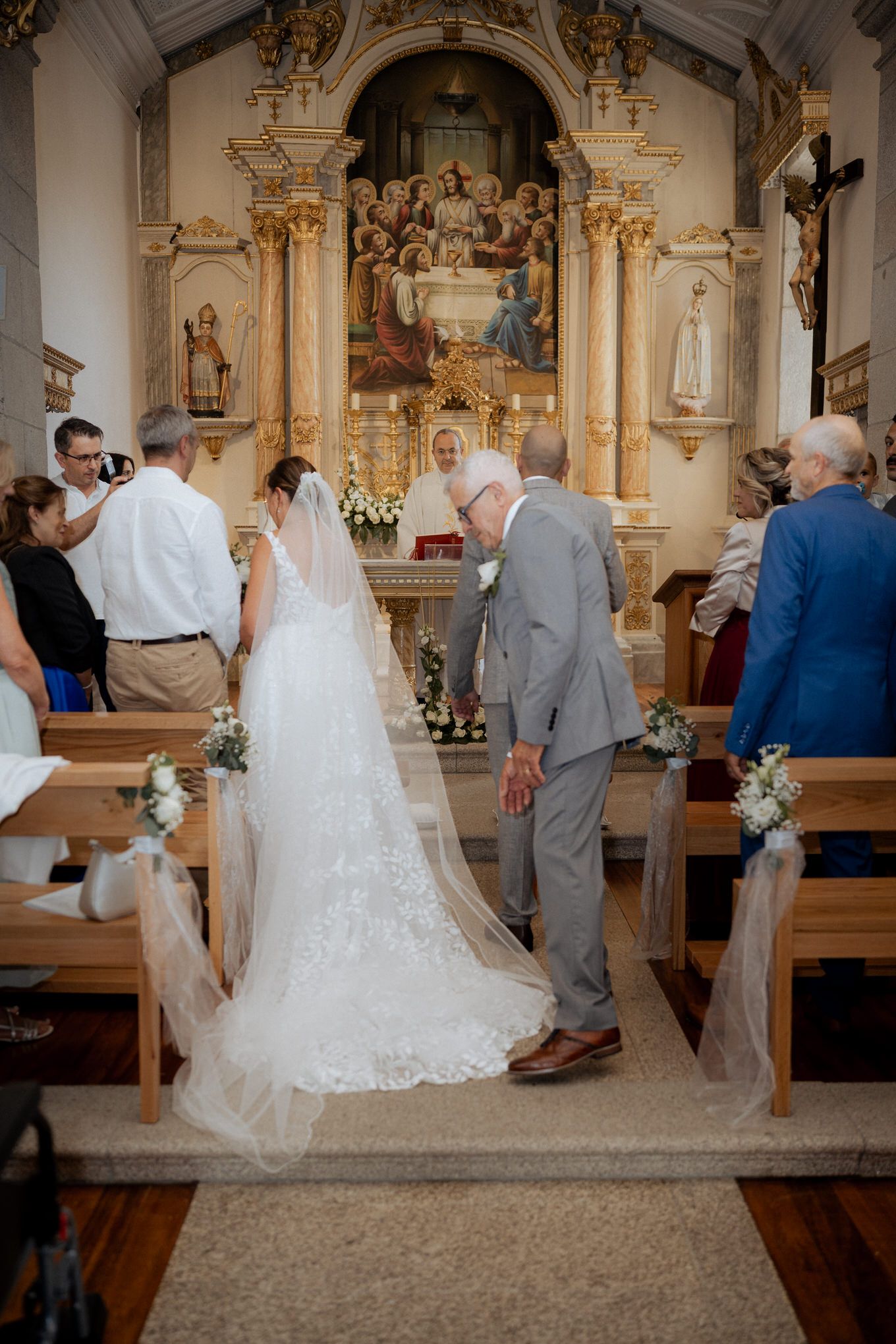 A wedding ceremony takes place in an ornate Catholic church with white floral decorations along the wooden pews.
