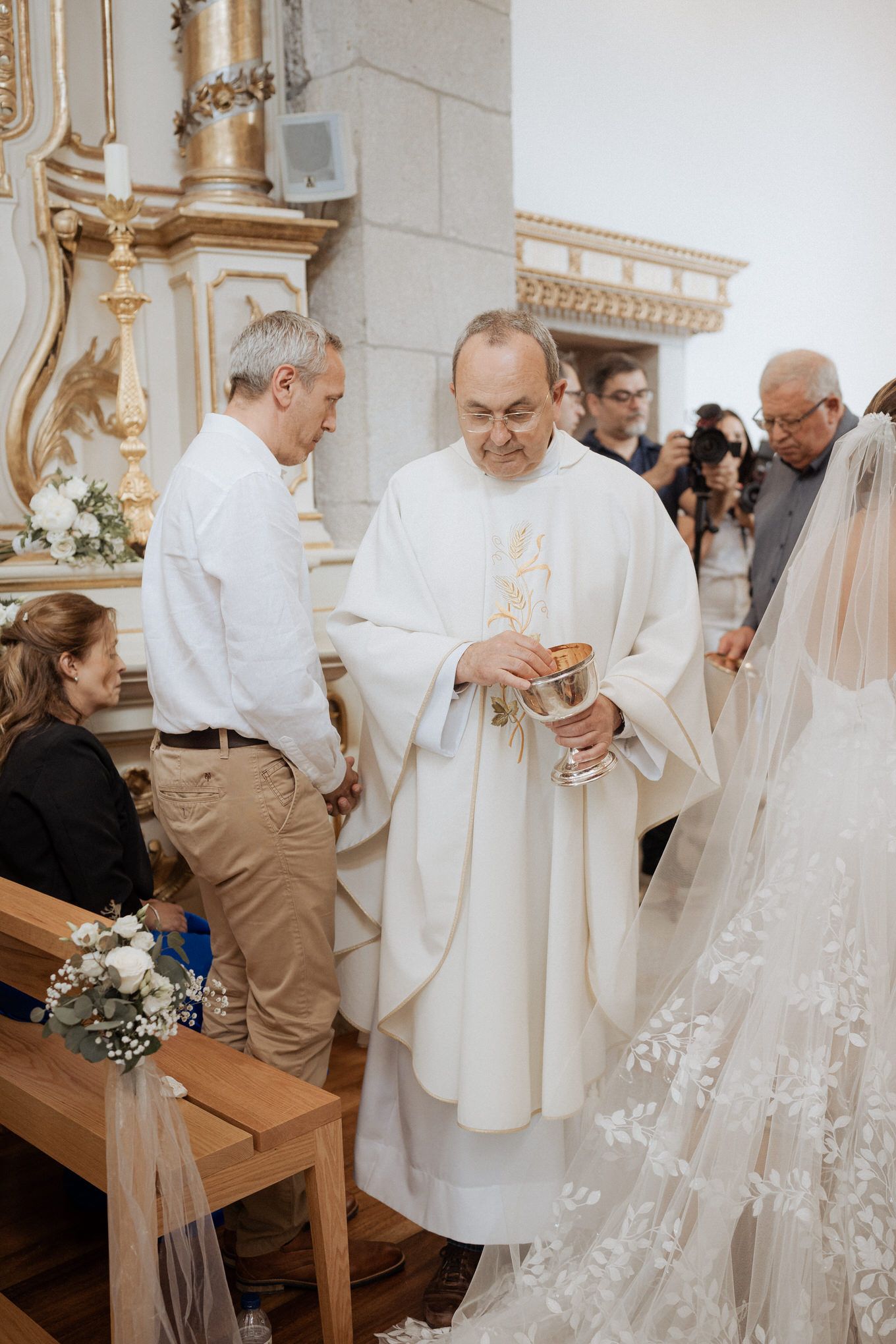 Priest in white robes stands near delicately laced wedding dress during a traditional church ceremony.