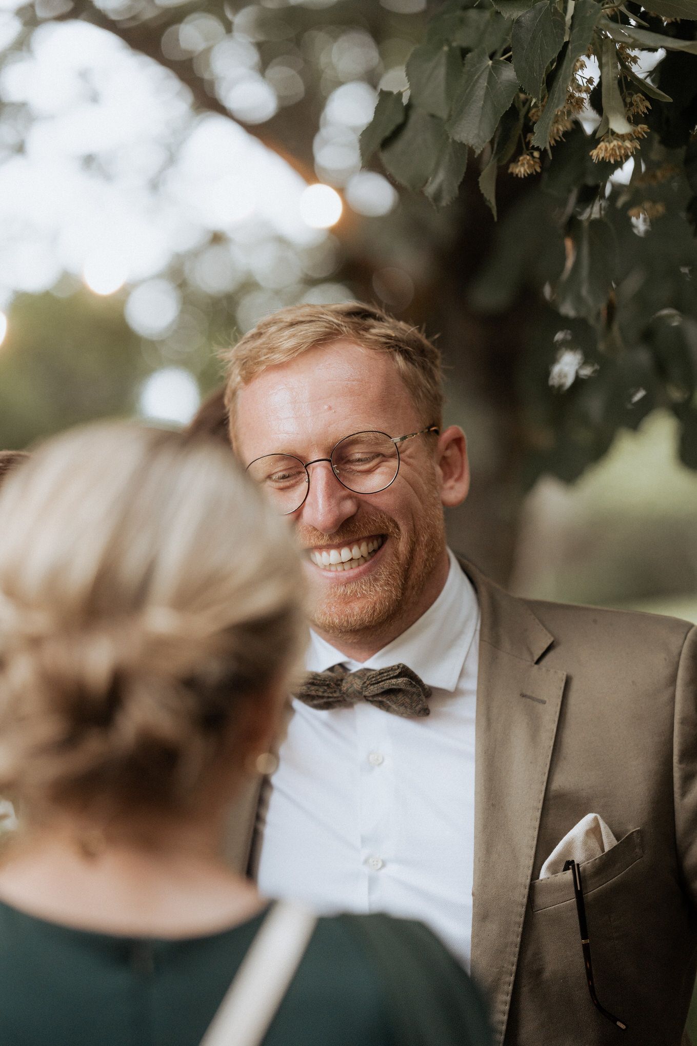 A person in glasses and a brown suit jacket with bow tie smiles warmly in an outdoor setting with bokeh lighting.