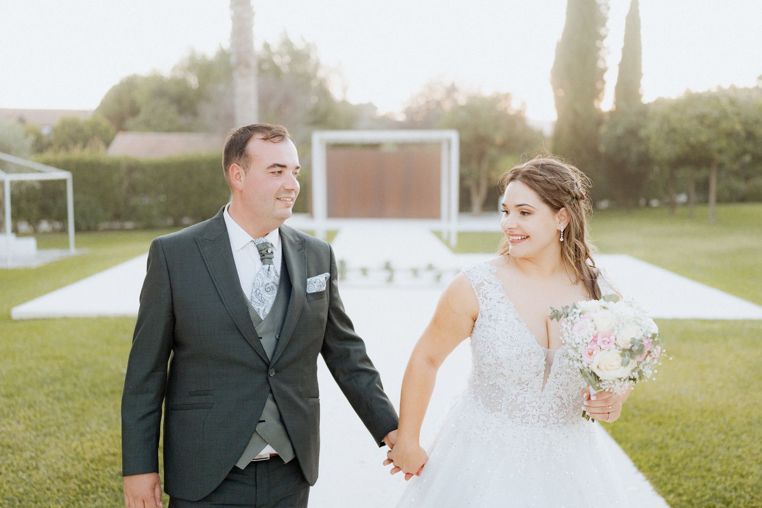 Happy newlyweds walk hand in hand on a grassy lawn with a white gown and dark suit on their wedding day.