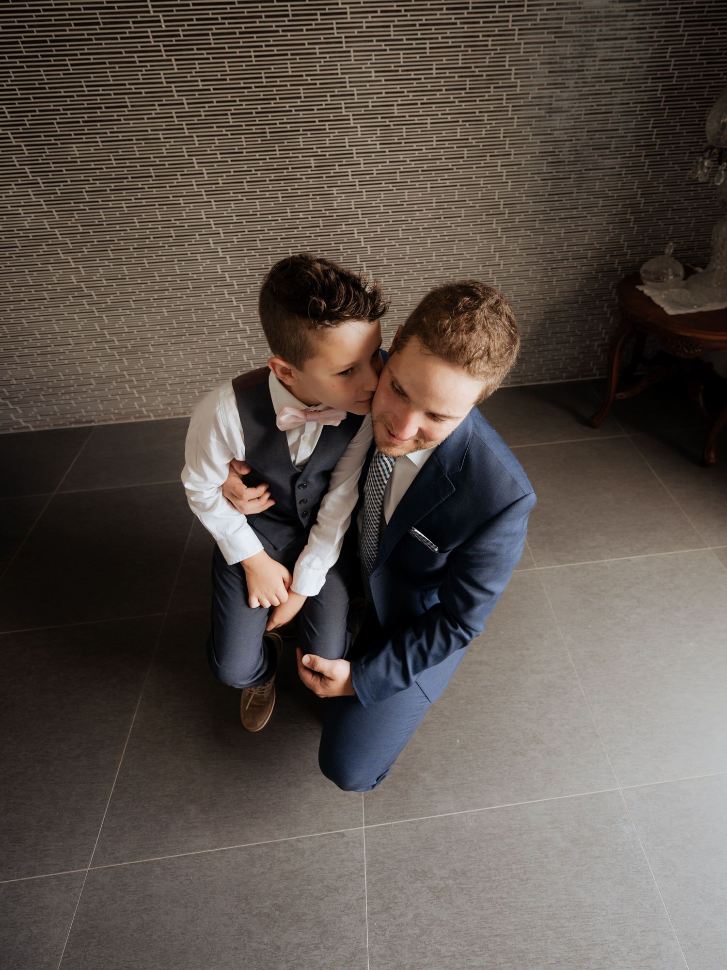 Two people in formal wear share an intimate moment against a textured grey wall.