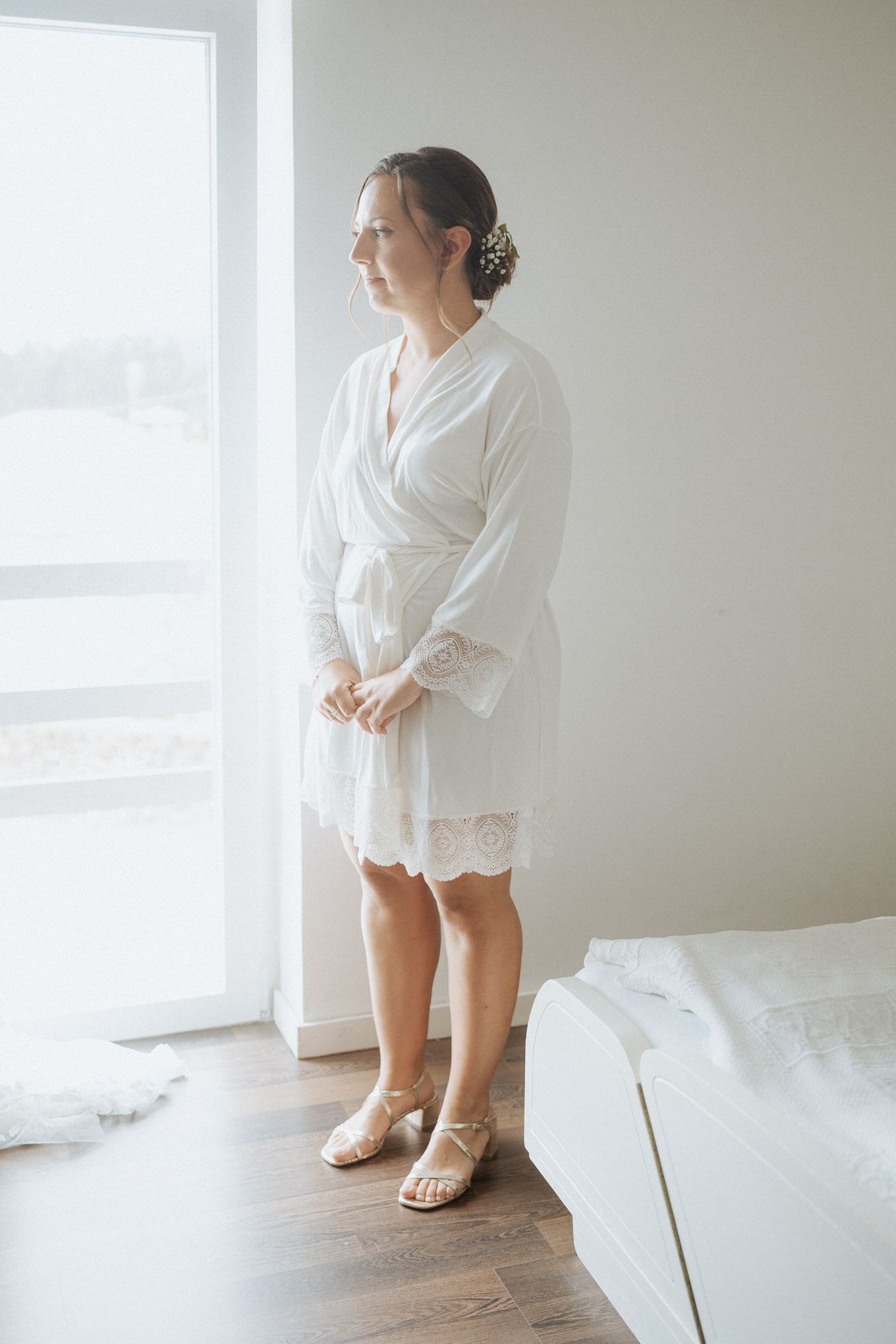 Standing by a bright window in a white lace-trimmed robe and sandals with hair elegantly styled in a bun.