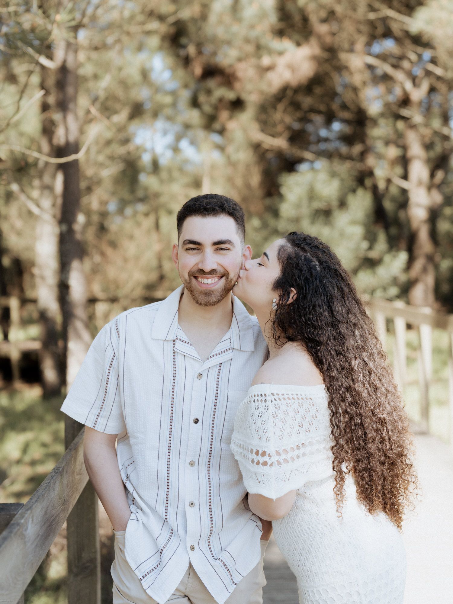 Um casal com trajes brancos a partilhar um momento doce juntos num cenário de floresta iluminada pelo sol.