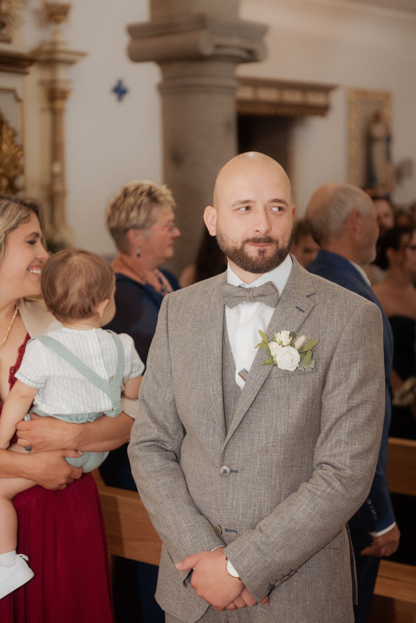 A groom in a gray suit with bowtie and boutonniere stands in a church during a wedding ceremony.