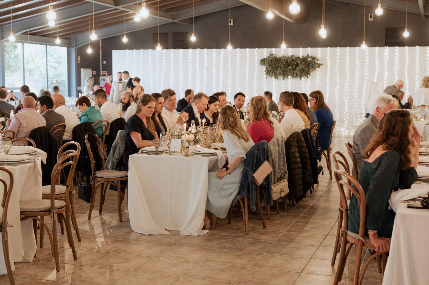 Wedding reception venue with guests seated at long tables under string lights and white drapes.
