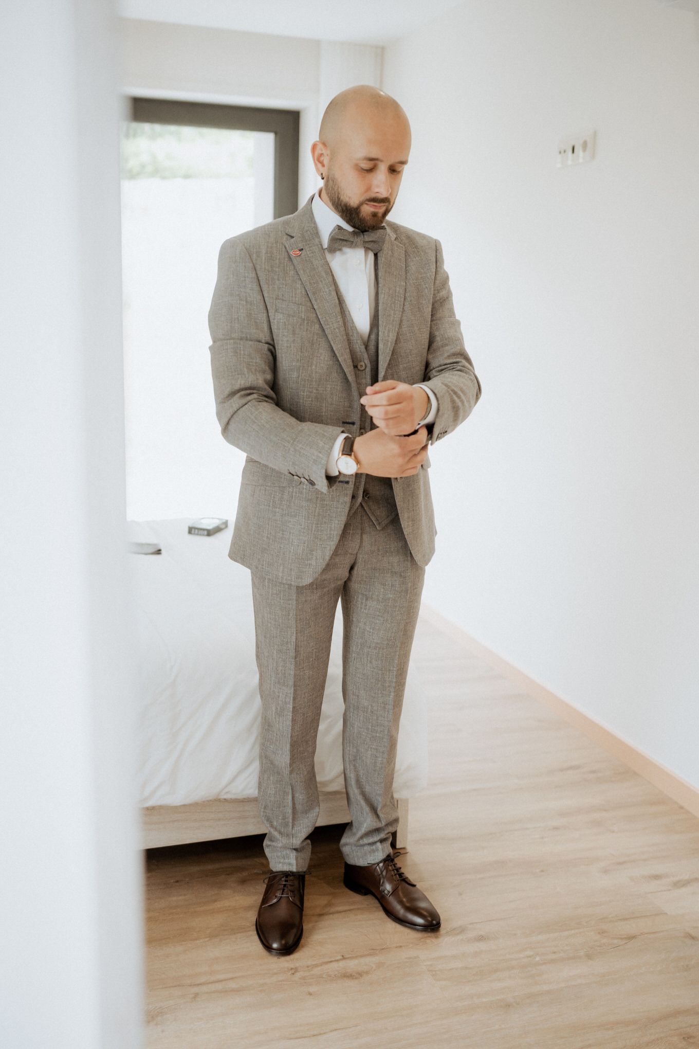 A person in a grey suit and bow tie adjusts their cuff while standing in a minimalist white room with hardwood floors.