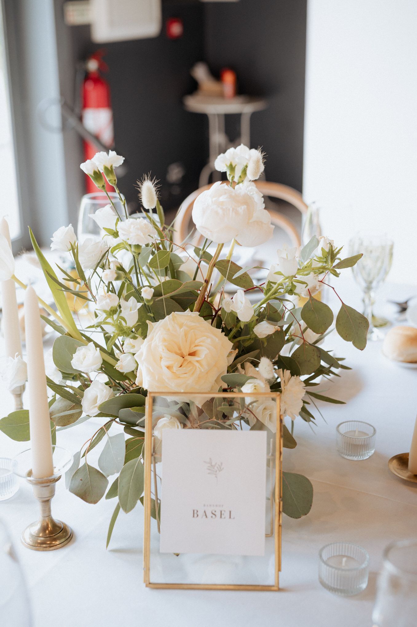 A romantic floral centerpiece with white flowers and greenery next to a gold-framed table number card.