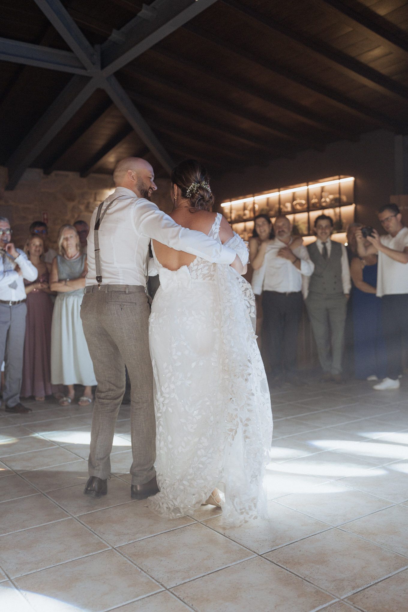 A couple shares their first dance at a rustic barn wedding reception while guests watch and clap from the sidelines.