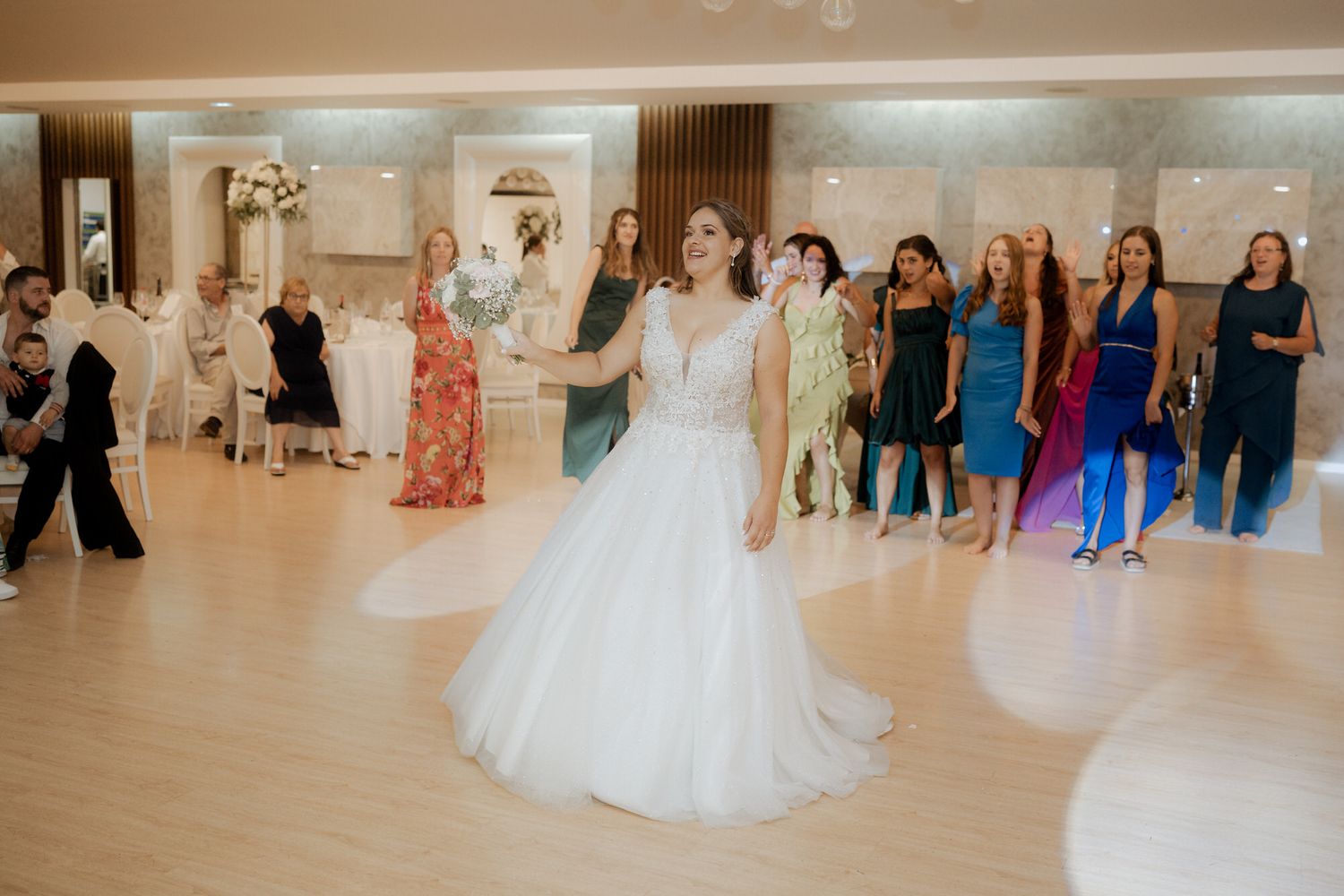 Wedding guests watch as a pair dances in a light-filled ballroom with hardwood floors during a reception celebration.