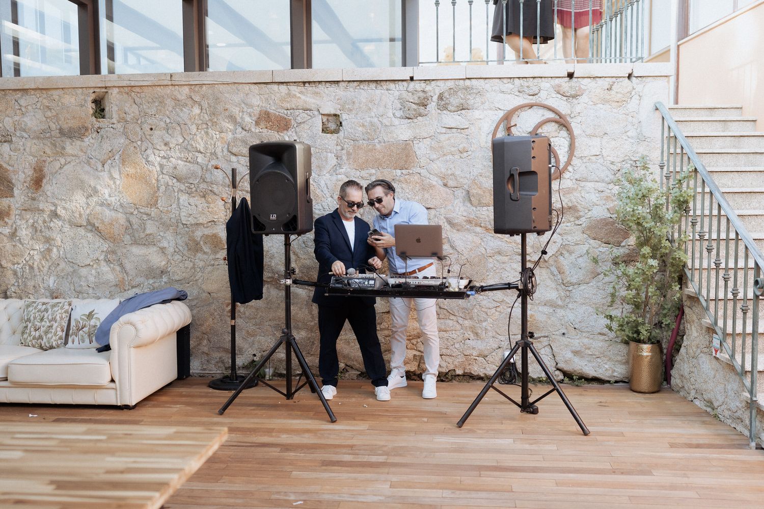 A DJ performs at a venue with professional speakers and equipment setup against a stone wall interior.