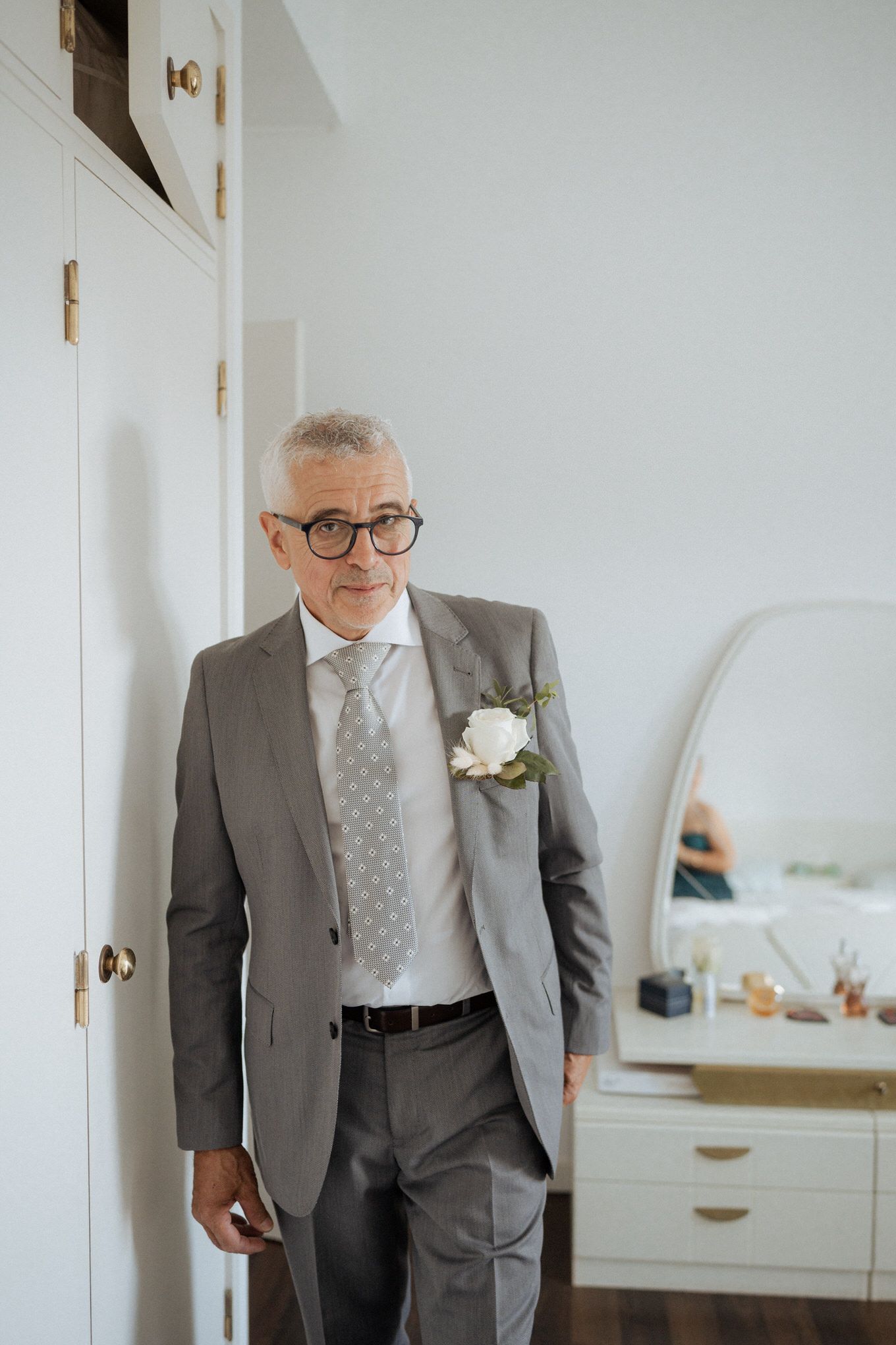 Well-dressed person in a grey suit and polka dot tie wearing glasses with a white boutonniere standing by a wall.