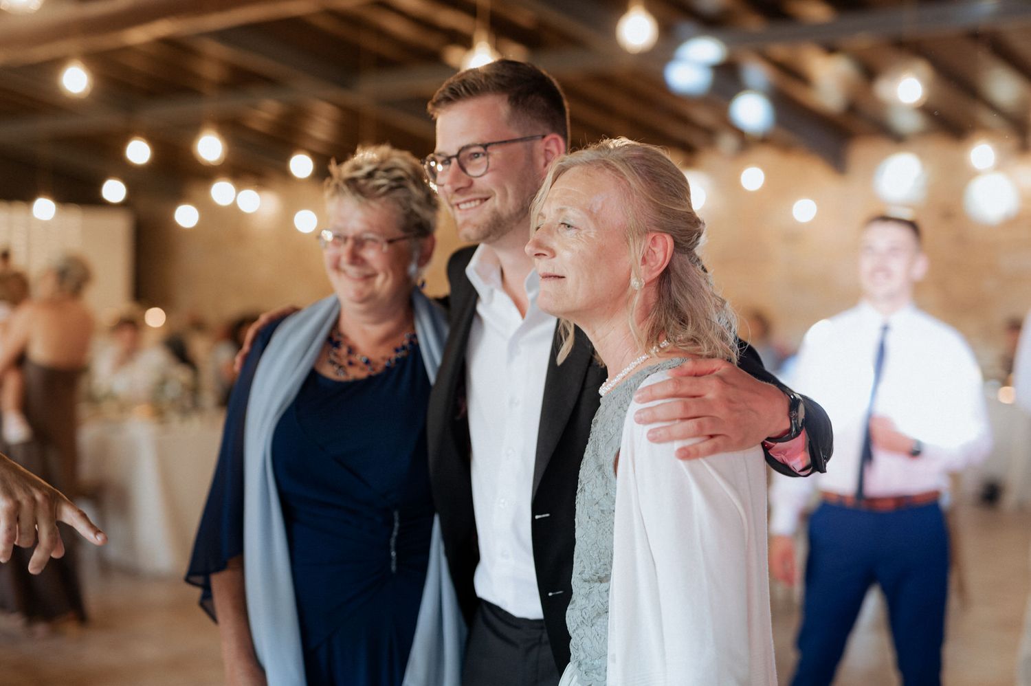 Three guests share an embrace under warm string lights at an indoor celebration venue.