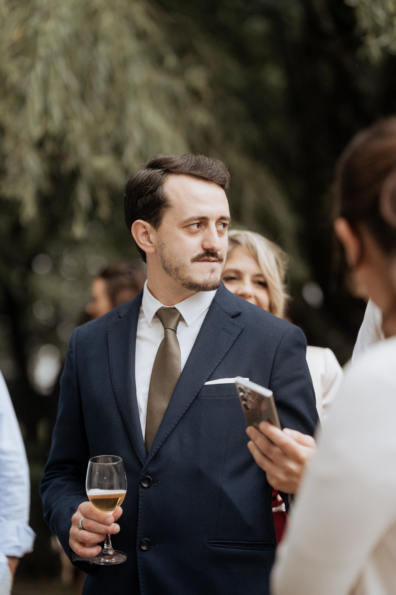 Wedding guest in navy suit holding champagne glass at outdoor reception.