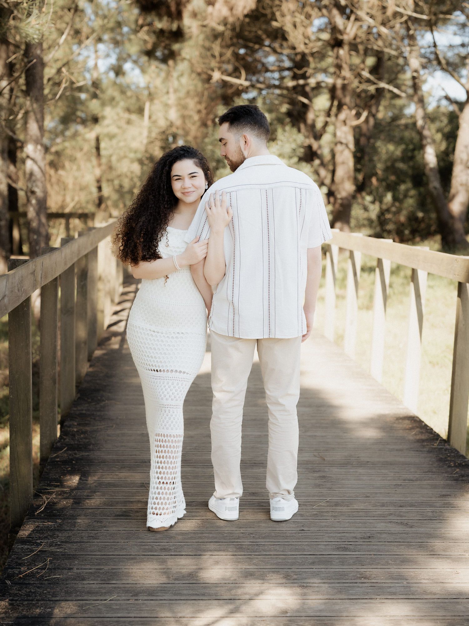 Um casal com trajes brancos iguais está junto num passeio de madeira rodeado por árvores.