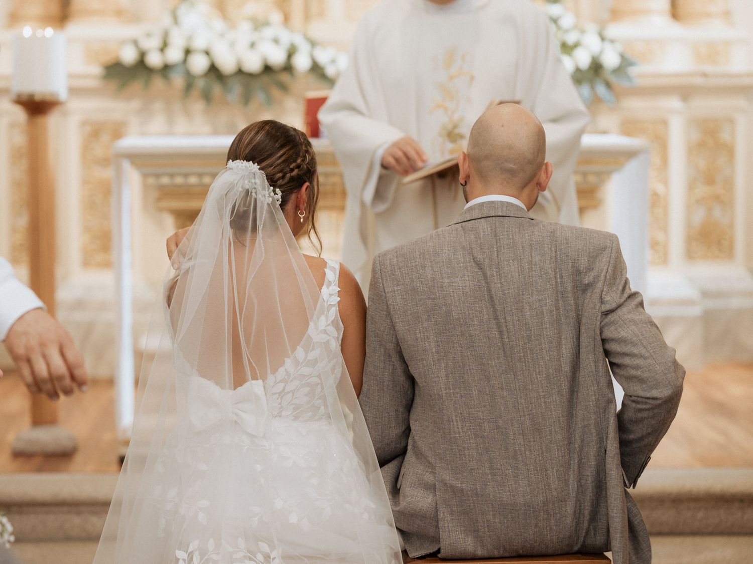 Bride in white lace gown and groom in grey suit stand at ornate church altar during wedding ceremony.