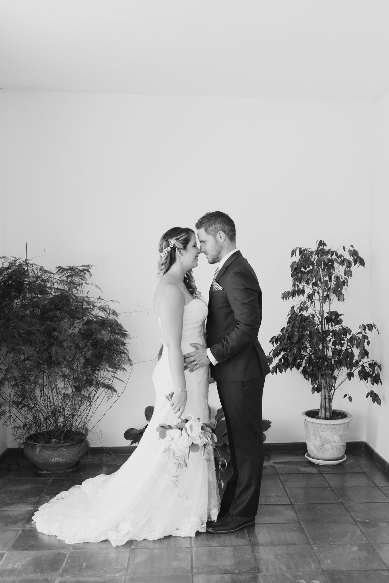 Black and white photo of newlyweds sharing an intimate moment against a wall with potted plants.