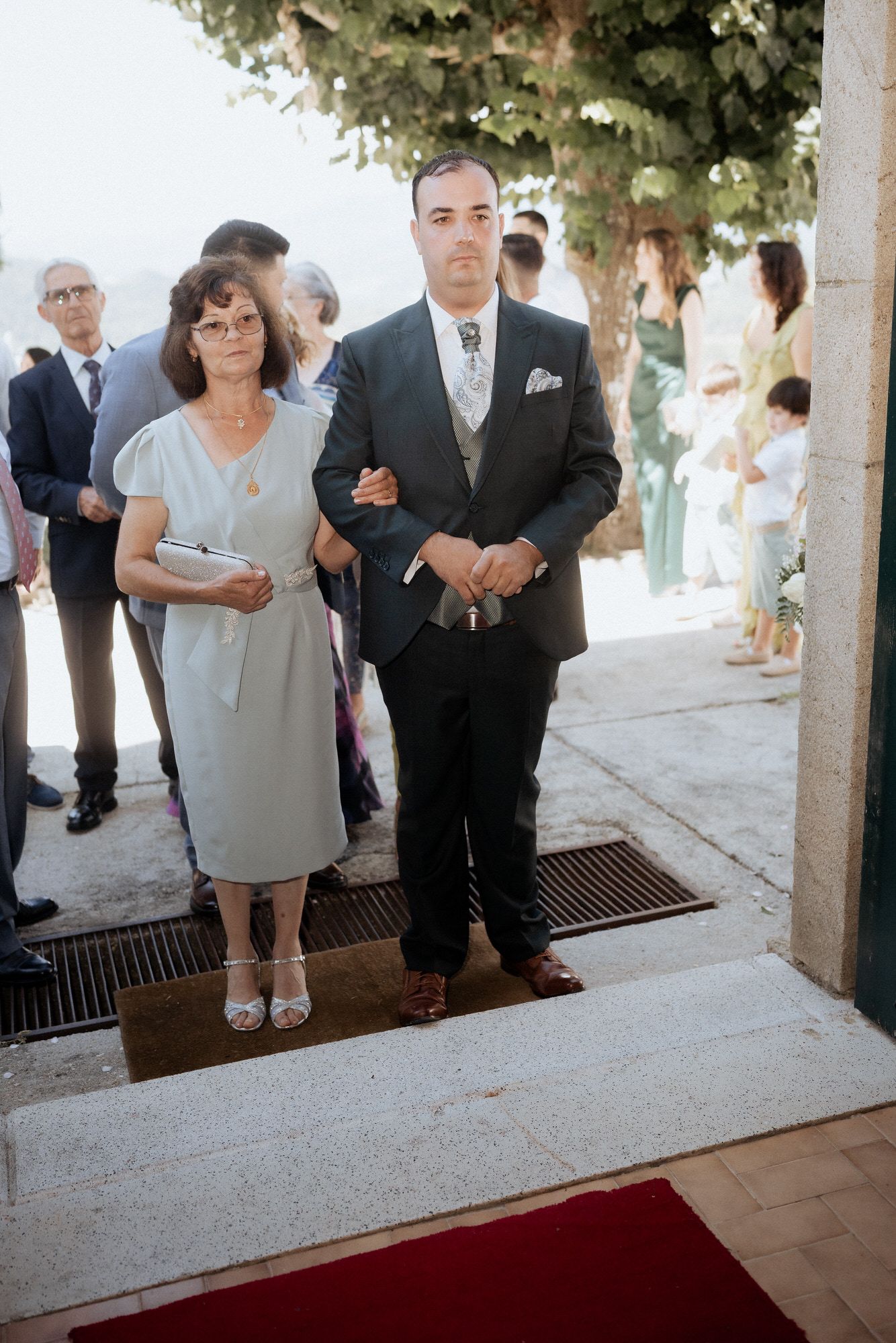 Guests stand along a tiled walkway with a red carpet at an outdoor ceremonial event.
