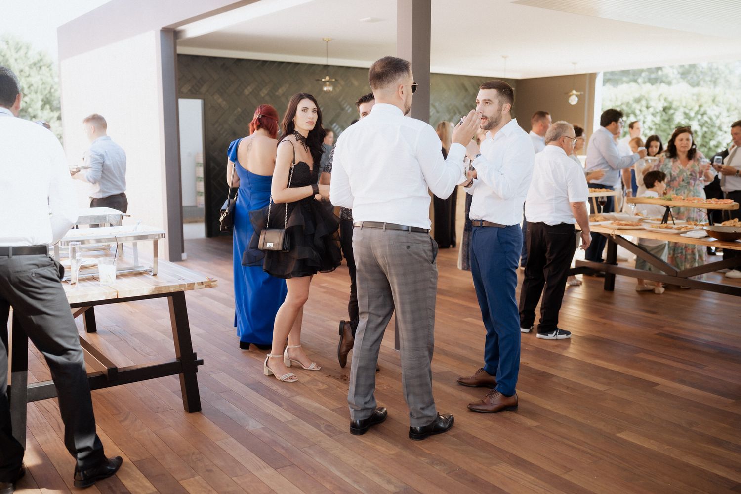 Guests mingle and socialize during an indoor event on a polished hardwood floor near buffet tables.