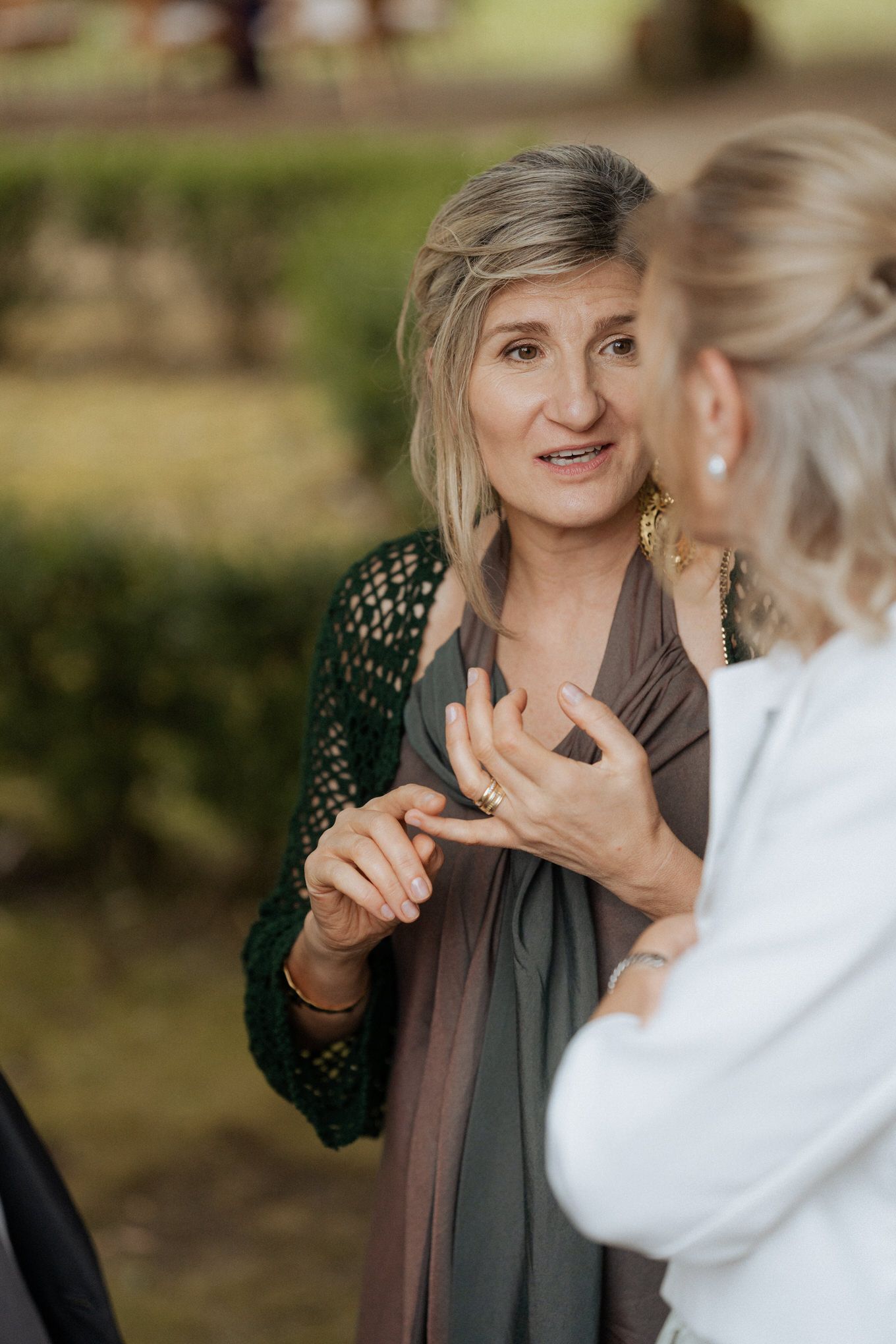 Two people engaged in animated conversation outdoors with one wearing a dark green crochet cardigan and neutral scarf.
