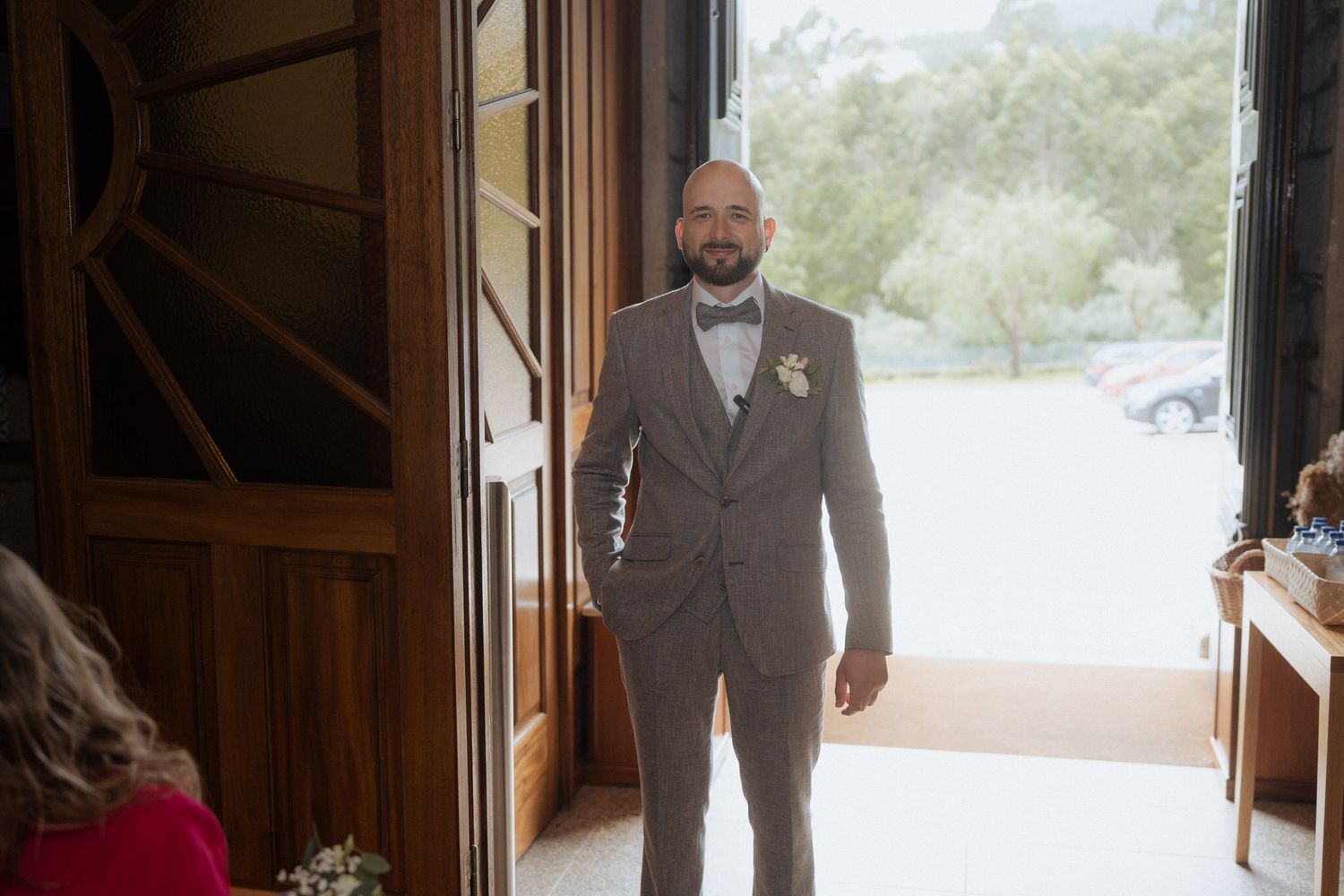 A groom wearing a brown suit stands in a doorway before his wedding ceremony.