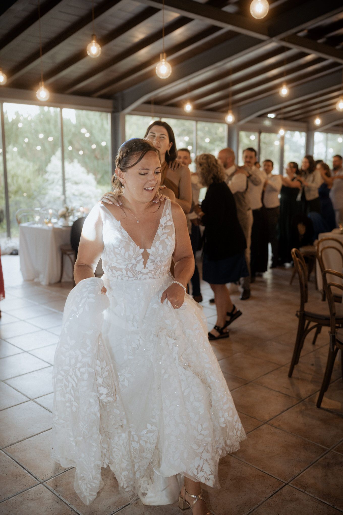 A bride in a detailed white lace wedding dress dances joyfully at a reception venue with exposed ceiling beams and lights.