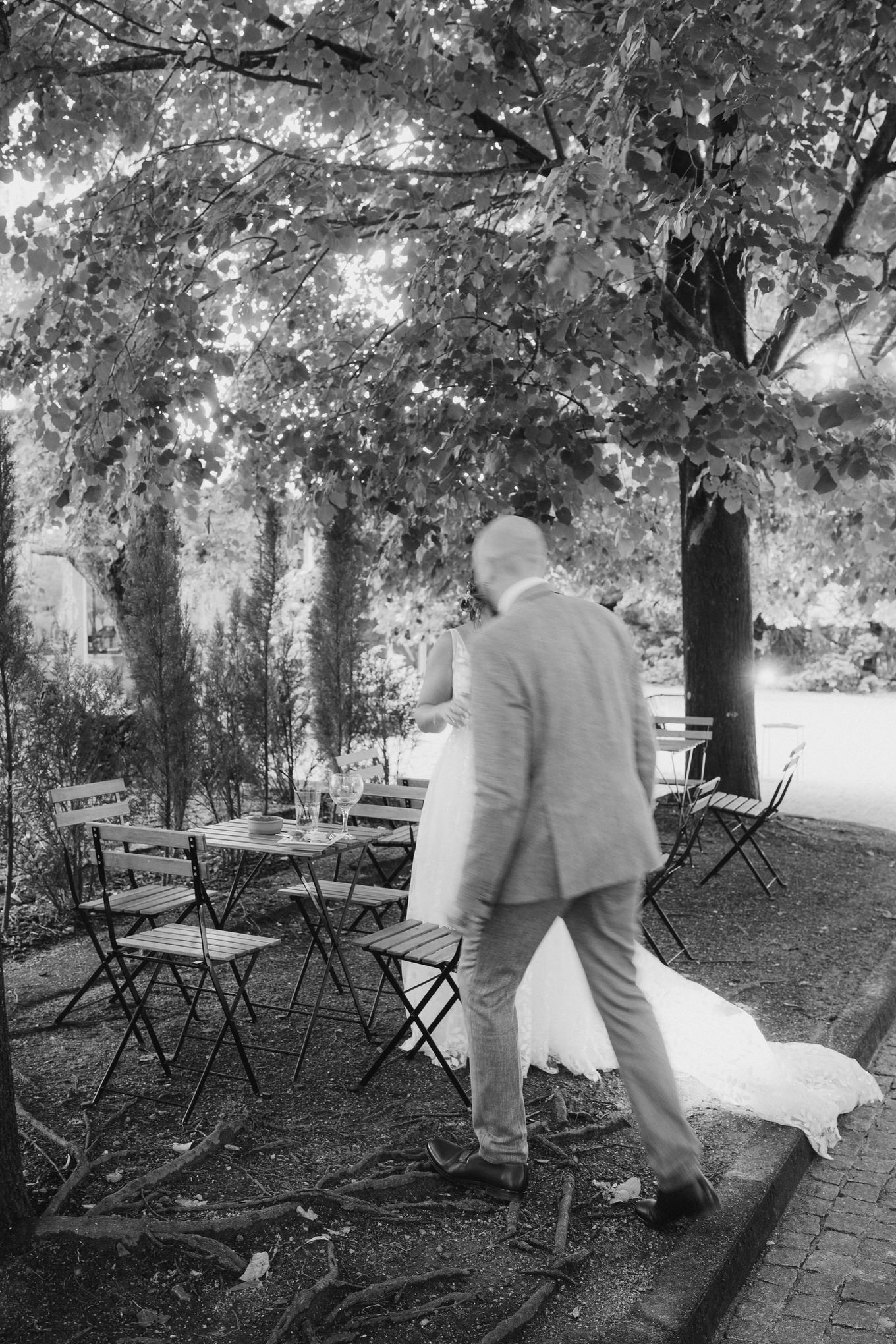 A person in a light colored suit walks among empty folding chairs under leafy trees in a black and white garden setting.