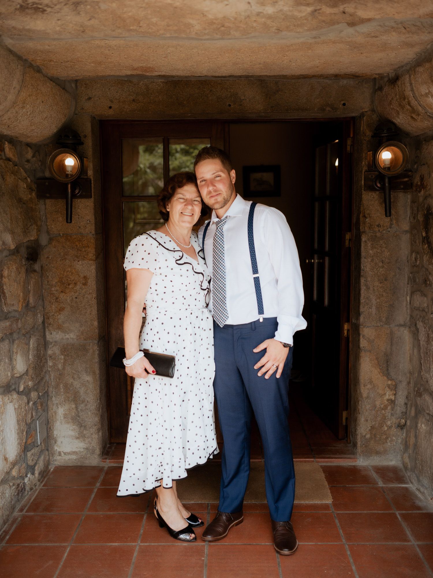 A couple stands in a stone archway entrance, dressed in polka dot dress and navy suspenders for a special occasion.