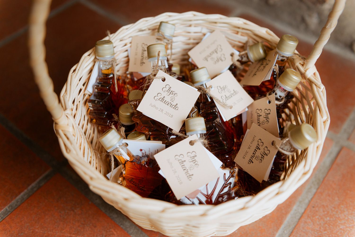 A wicker basket filled with homemade jam jars decorated with rustic labels and fabric tops on terracotta tile floor.