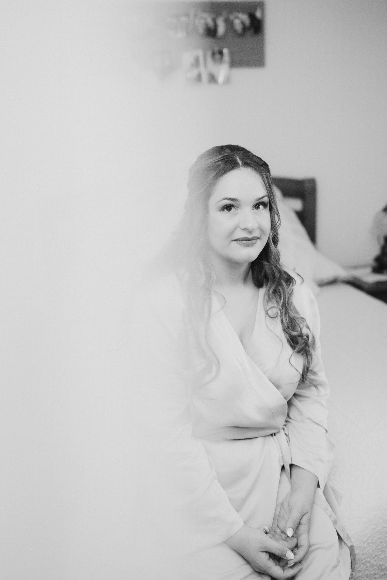 Black and white portrait of someone in a light-colored wrap dress sitting and posing against a plain wall.