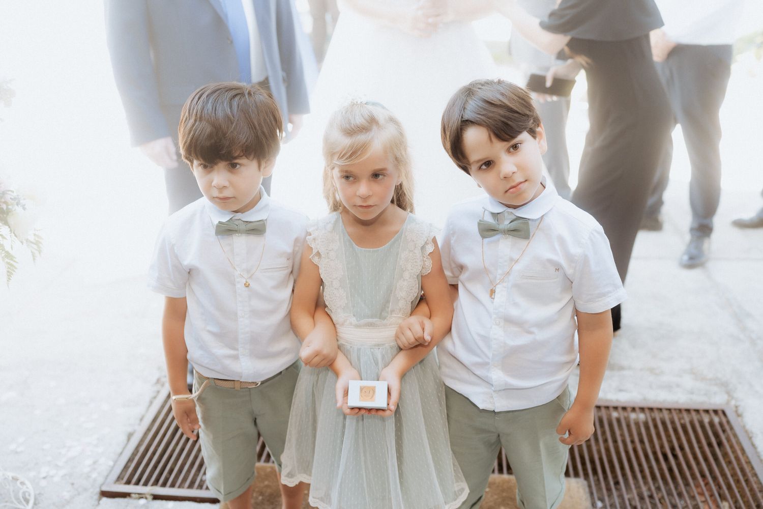 Three young children dressed formally in white shirts and bowties for a special occasion stand together on a sidewalk grate.