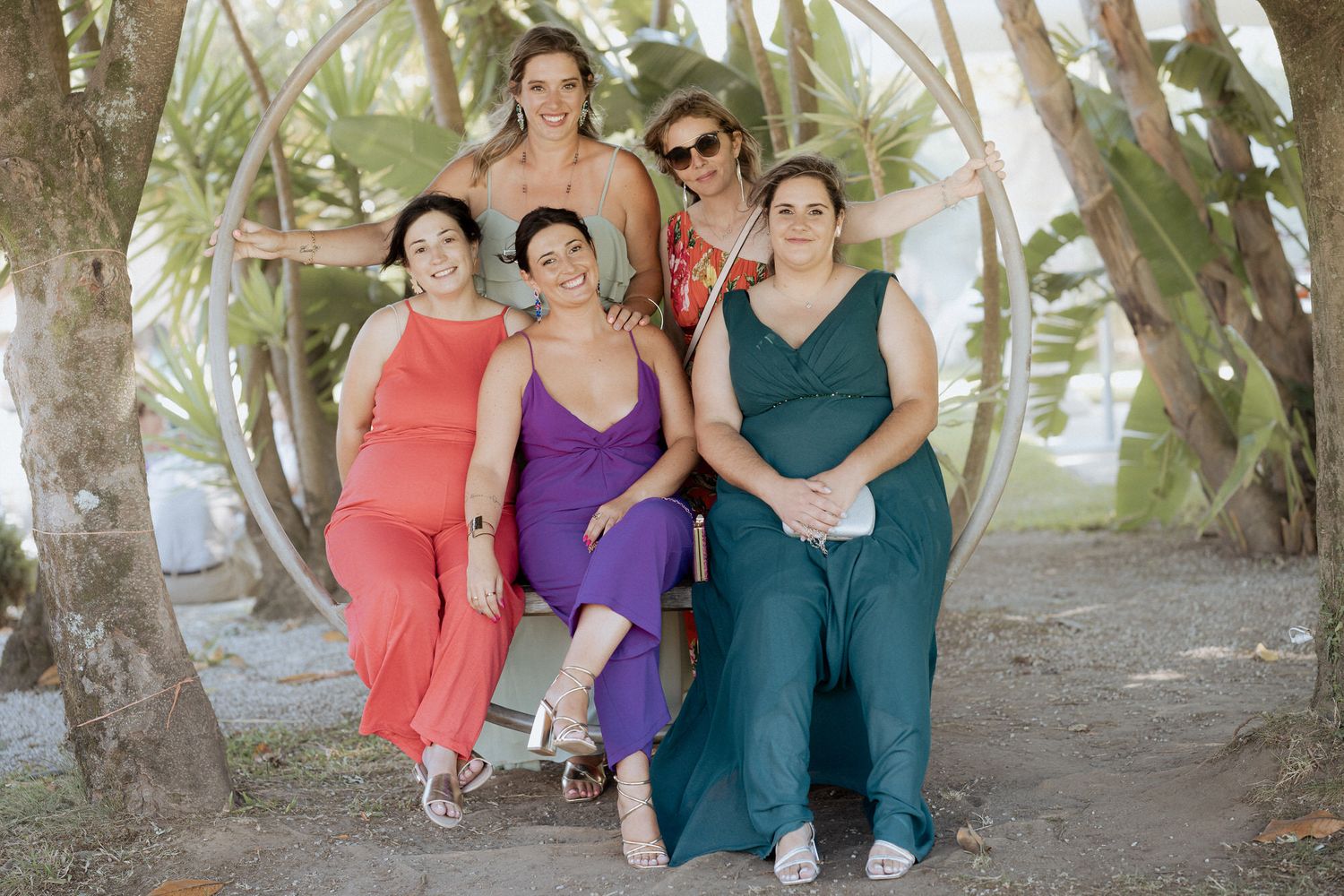 Four people wearing colorful jumpsuits in coral, purple and teal pose together outdoors among palm trees on a sunny day.