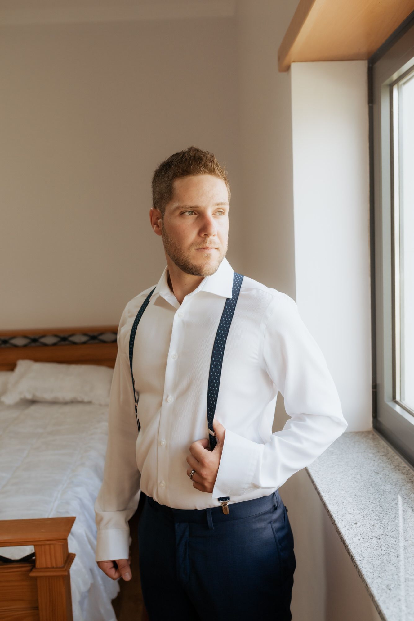 A person in a crisp white dress shirt and navy suspenders stands pensively by a window in a bedroom setting.
