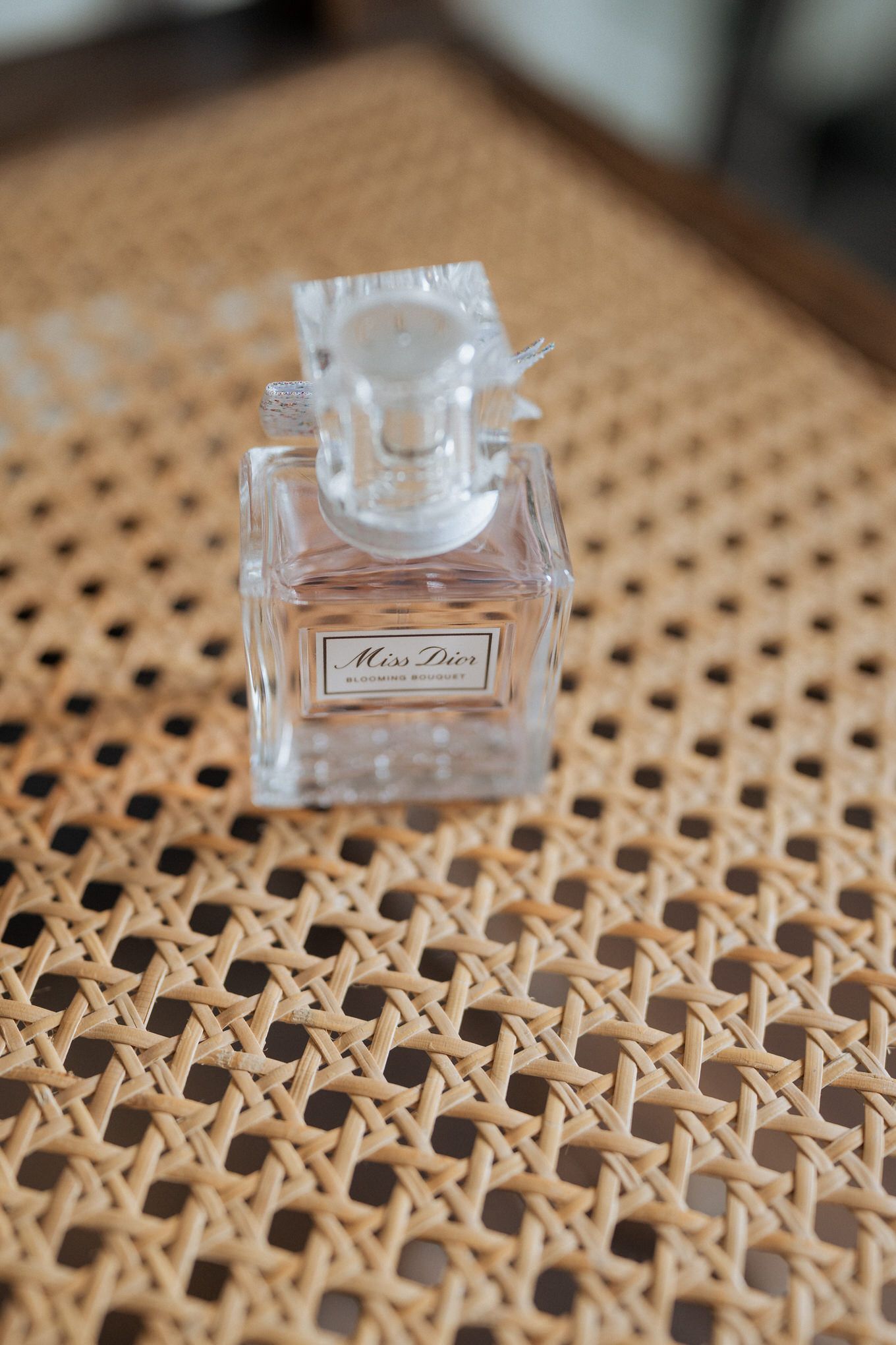 A small glass bottle of Miss Dior perfume sits on a textured woven rattan surface.
