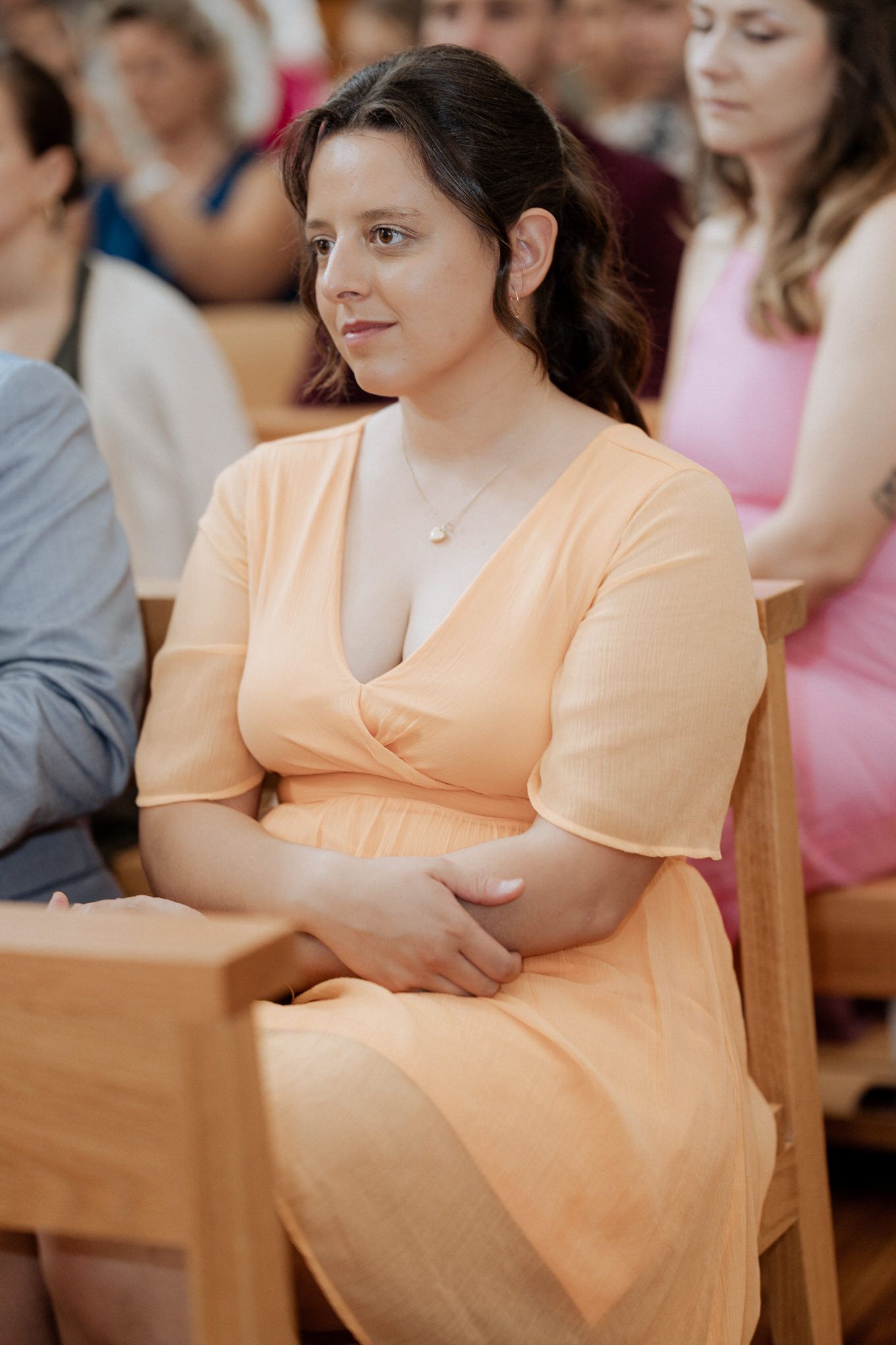 Guest in a peach wrap dress sits attentively during a ceremony in a wooden pew.