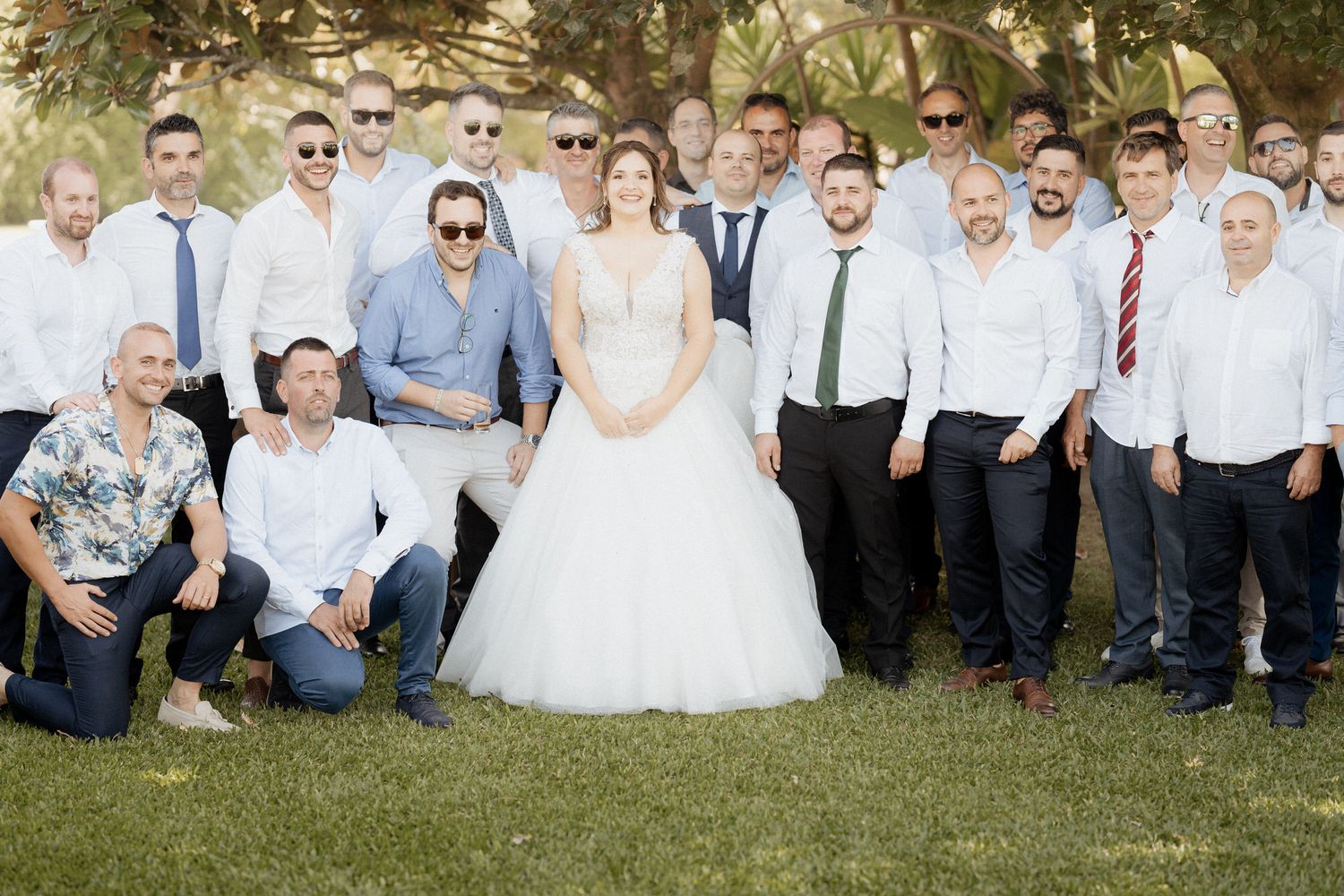 A group of wedding party members gather outdoors on grass for a formal photo in light and dark attire.