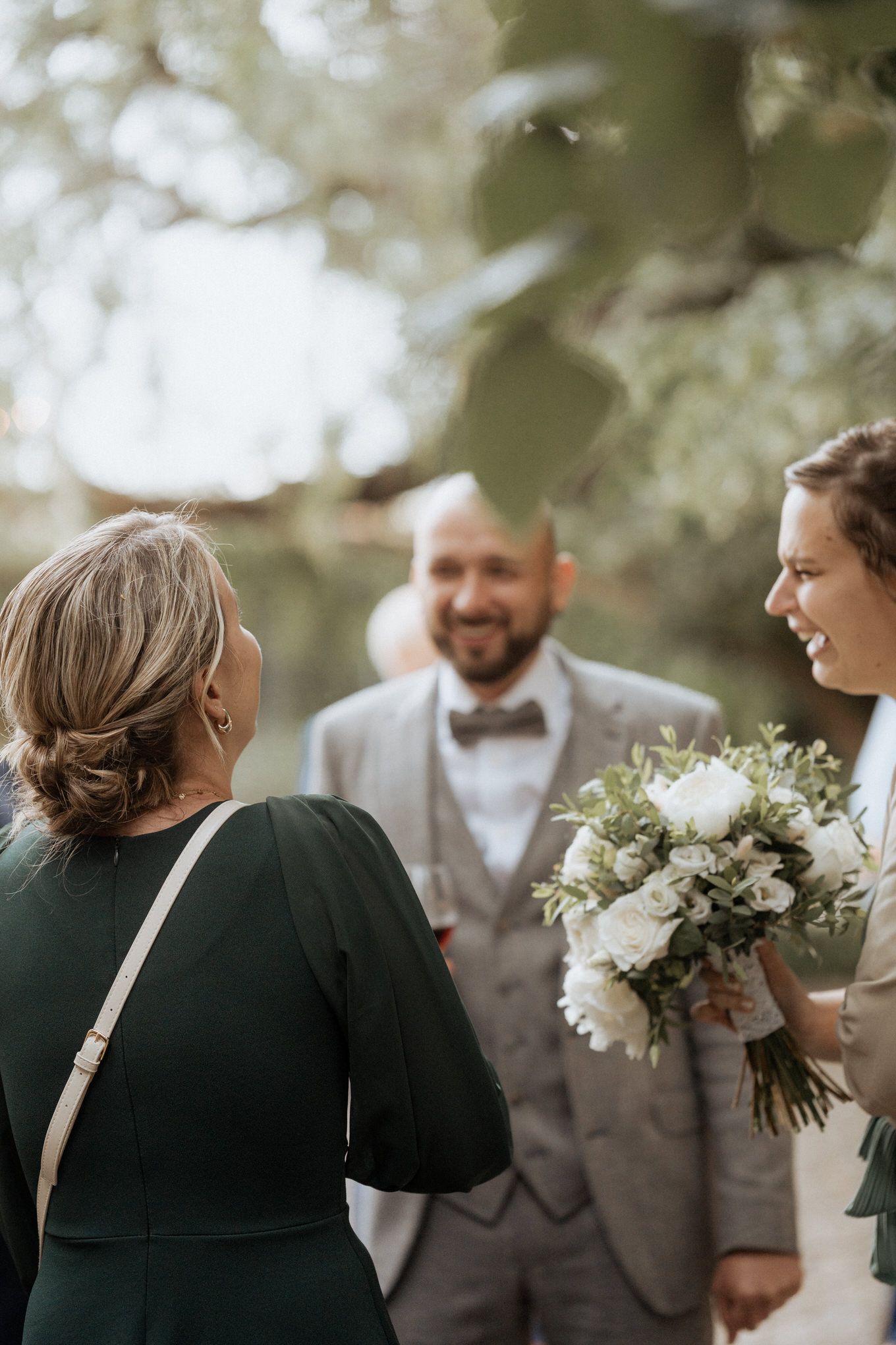 Intimate outdoor wedding ceremony shows a happy couple and officiant under blooming trees with a white floral bouquet.