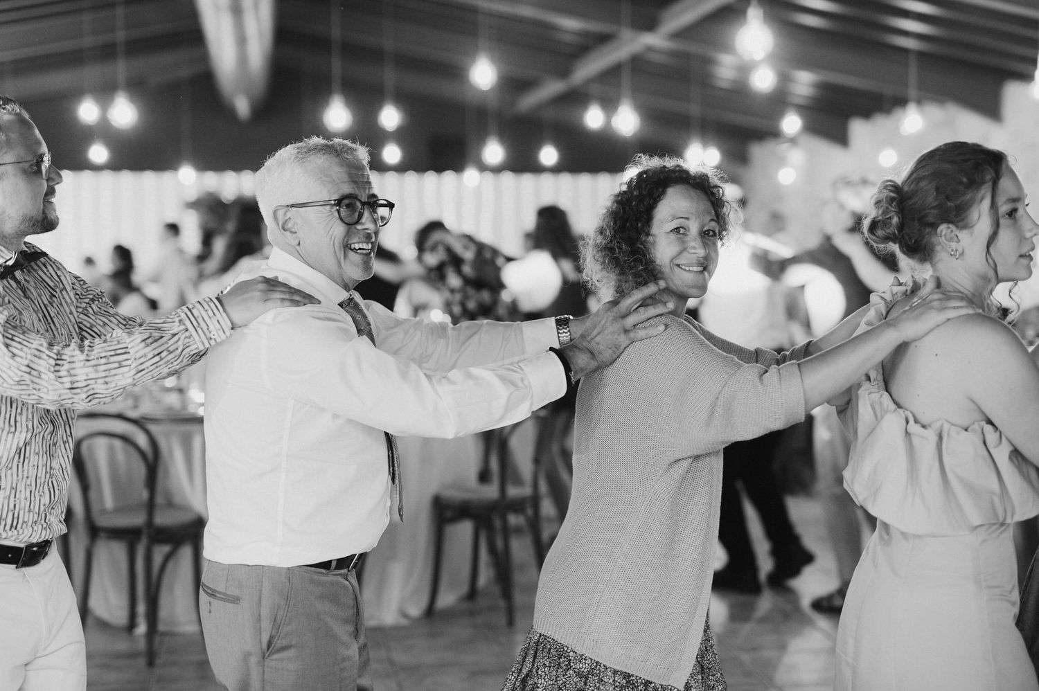 Wedding guests perform a line dance in black and white at an indoor reception with pendant lighting.