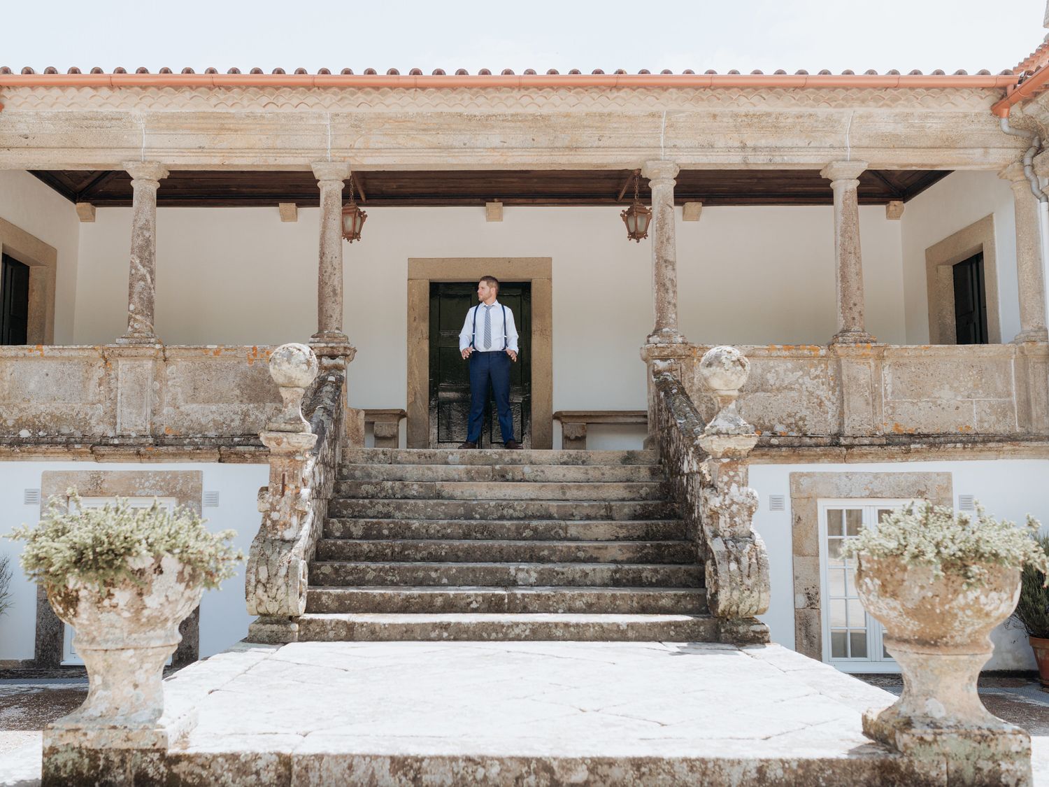 A figure stands at the top of white stone steps leading to an elegant colonial-style porch with decorative columns and planters.