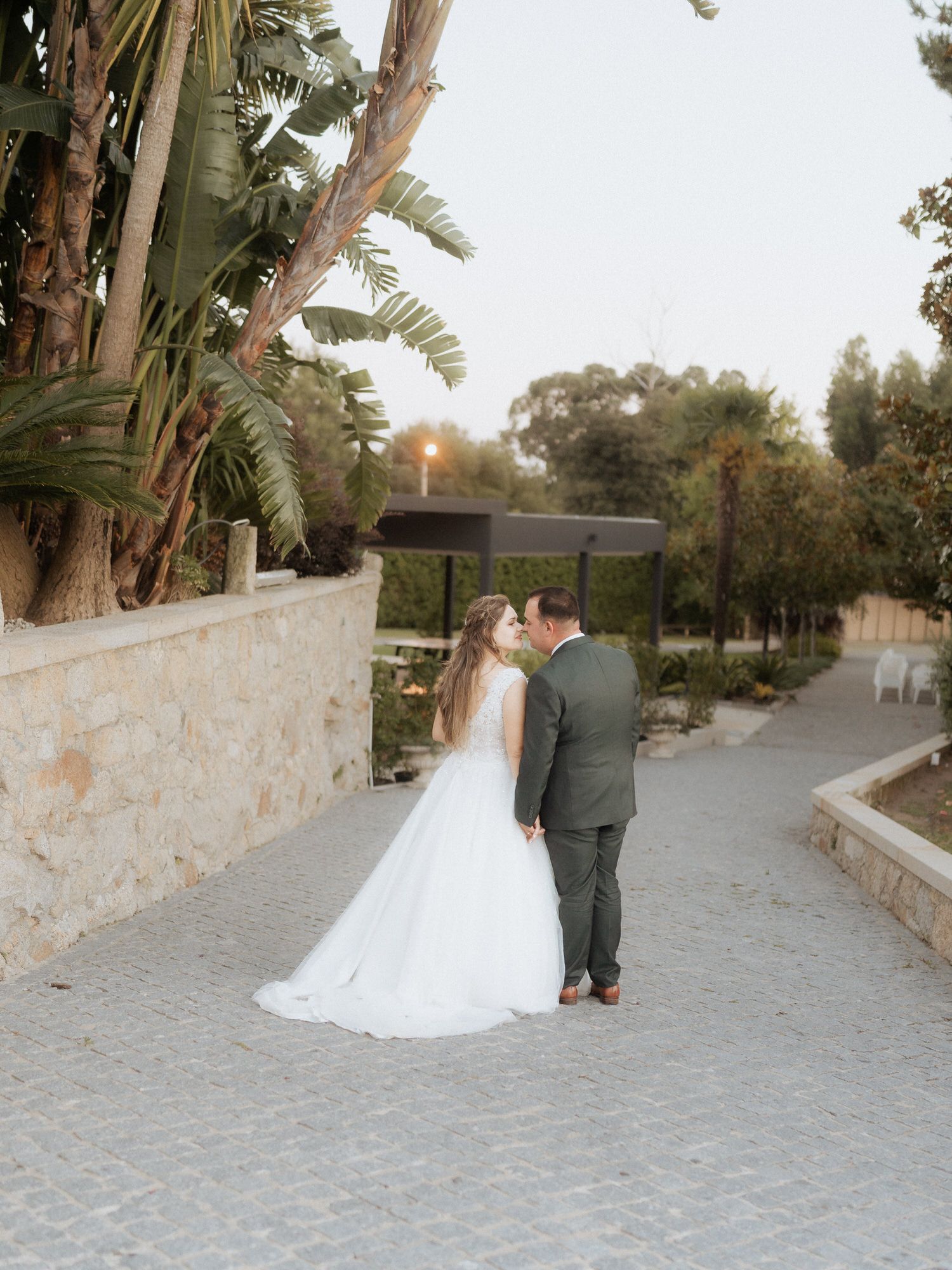 Romantic couple in wedding attire embracing on a stone path with palm trees and lush greenery in the background.