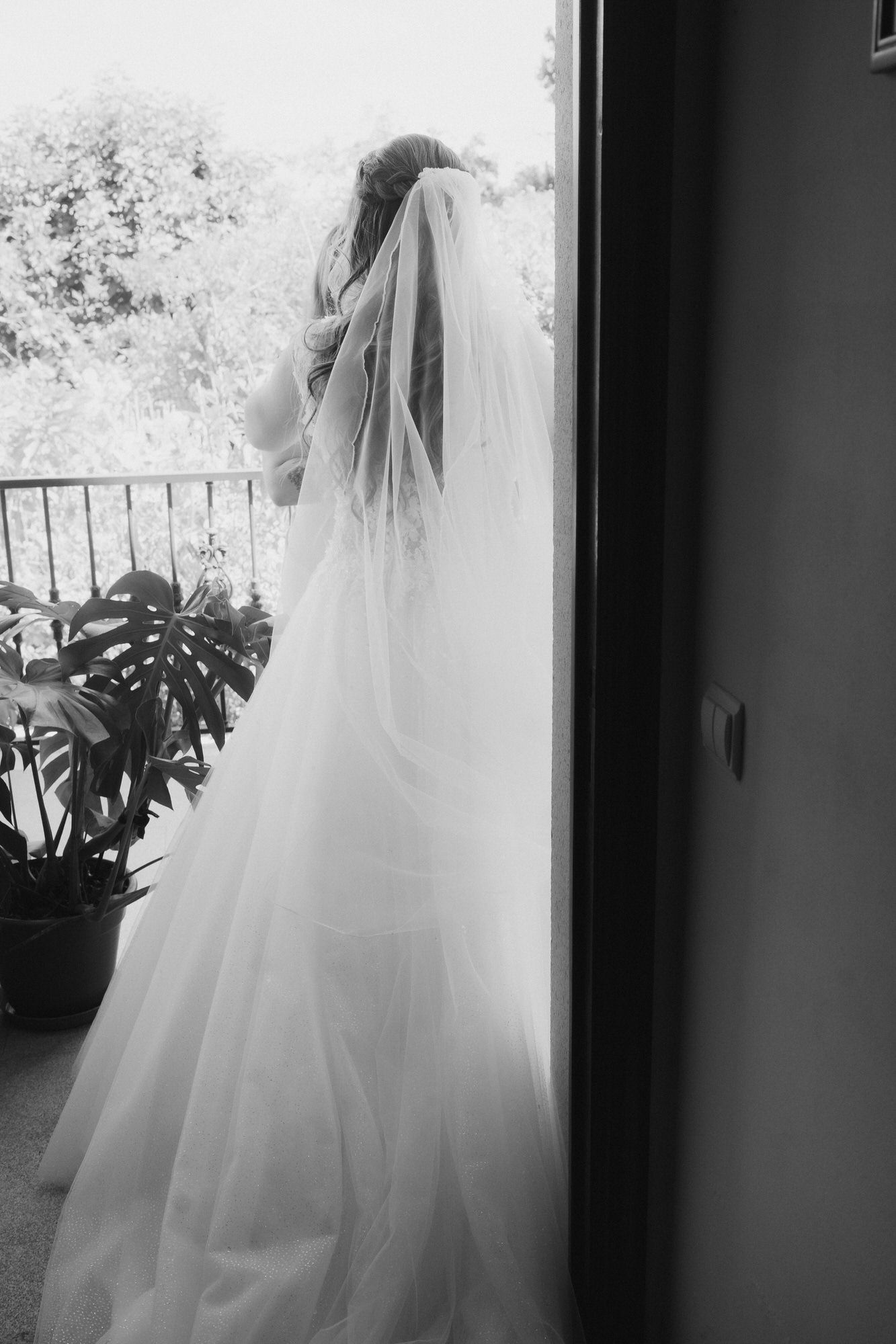 Black and white photo of a bride in a flowing veil and gown standing by a railing on a balcony.