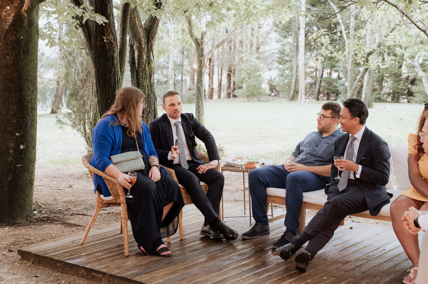 A group of people having a casual conversation while seated on chairs on a wooden deck surrounded by trees.