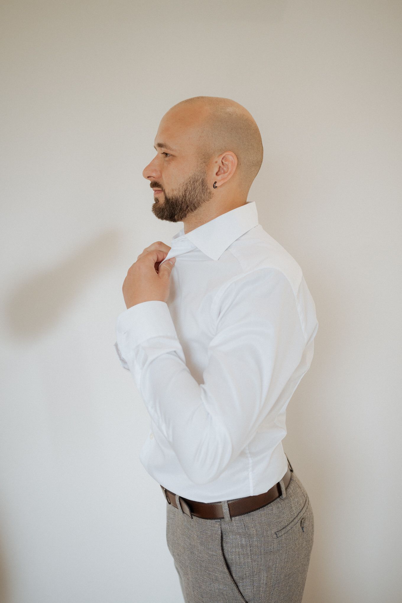 A professional in formal attire adjusts their collar while standing in profile against a plain wall.