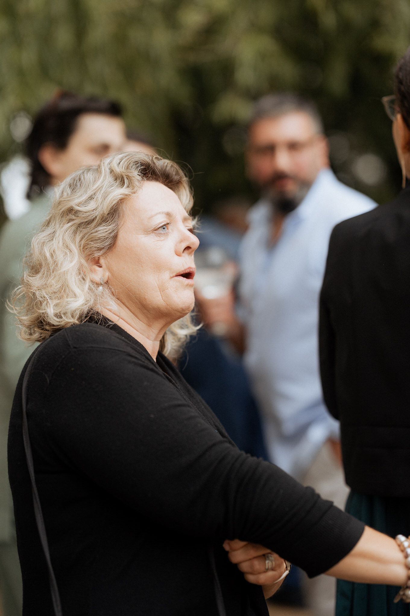 A person in a black sweater gestures animatedly while speaking outdoors with others visible in the background.