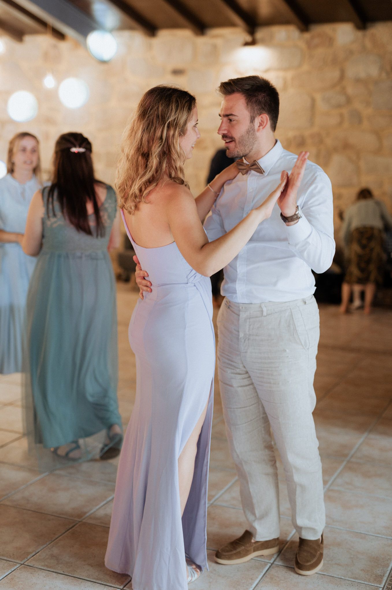 A couple dances intimately at a wedding reception in a rustic stone venue while bridesmaid in blue dress watches.