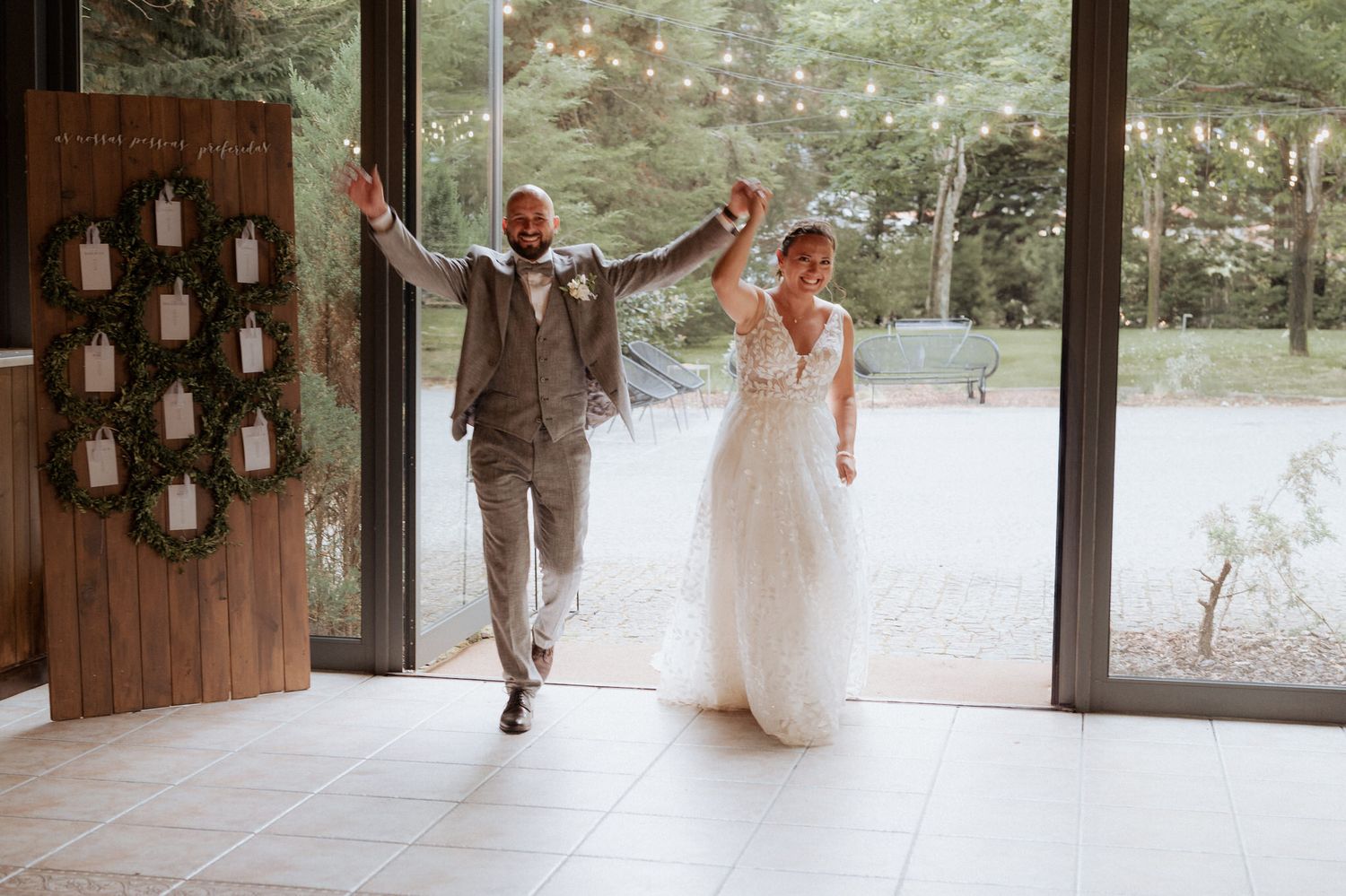 Joyful newlyweds celebrate while entering a rustic indoor venue with hanging lanterns and large glass windows.